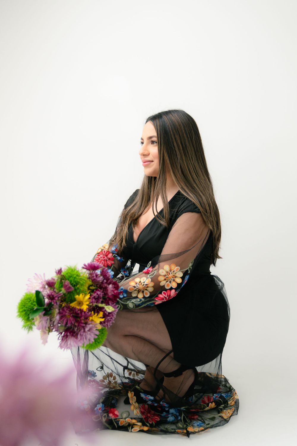 A person in a black dress kneeling on a floral cloth while holding a vibrant bouquet of colorful flowers.