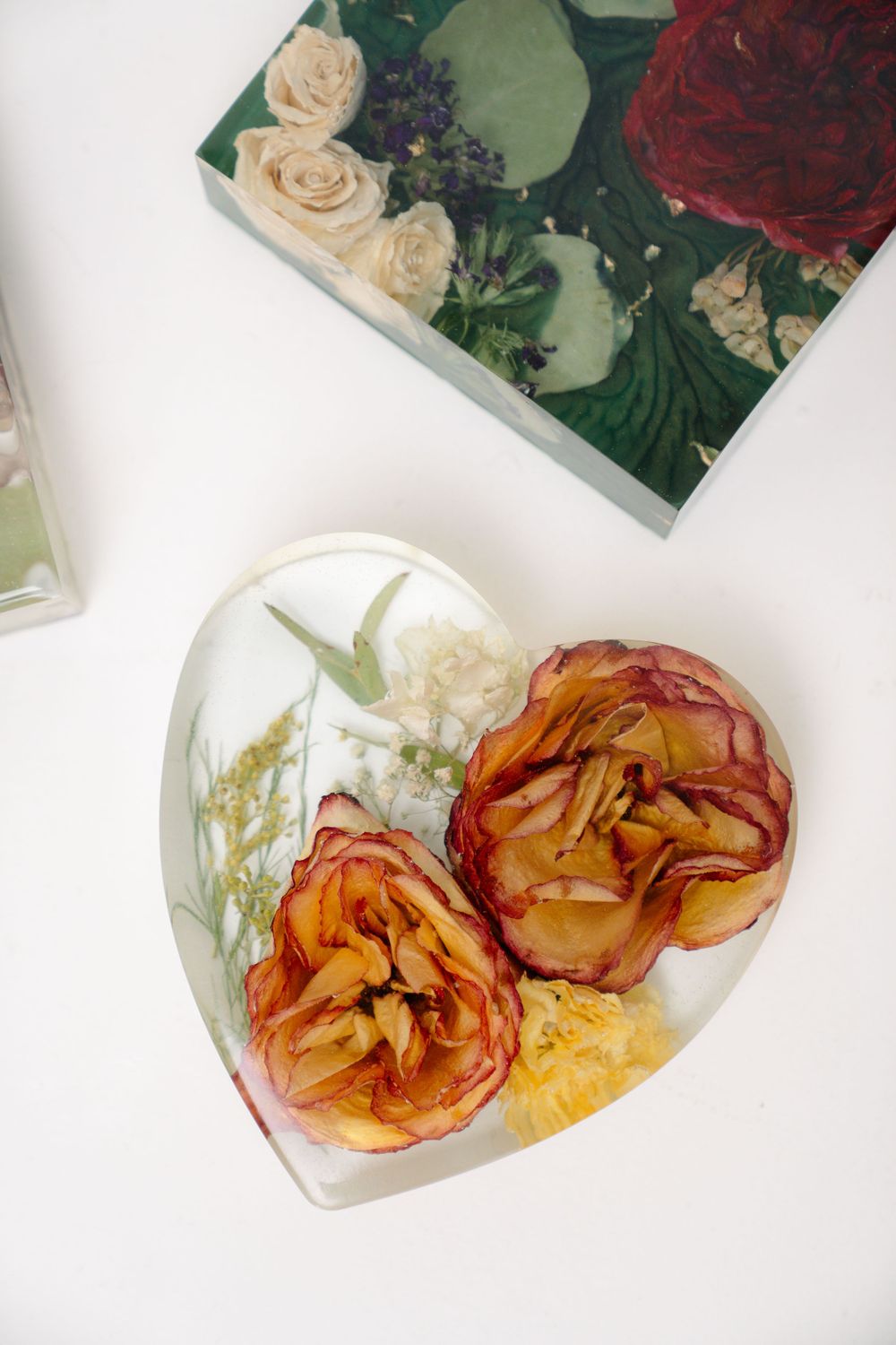 Dried orange roses preserved in a heart-shaped resin container with botanical elements on a white surface.