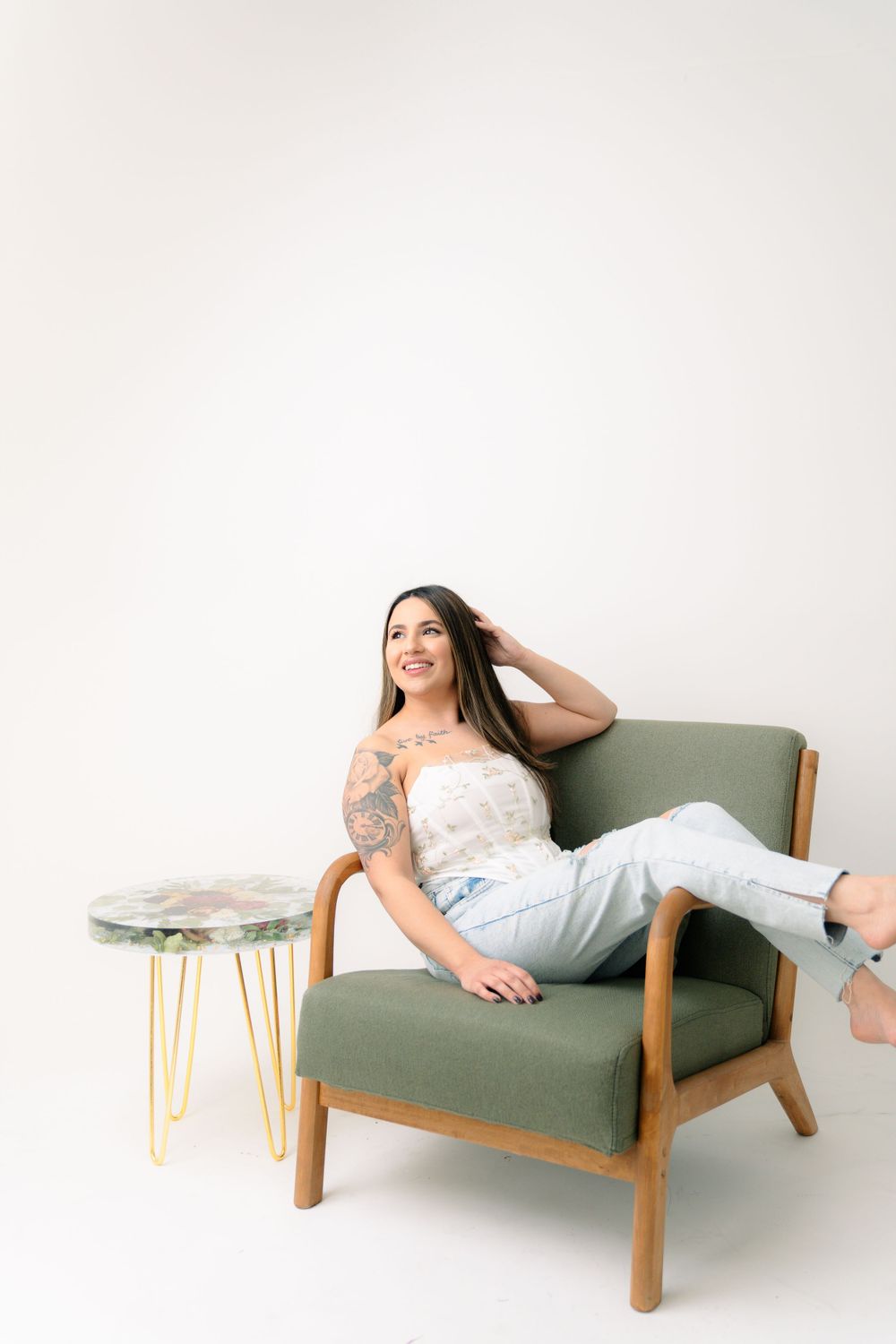 A woman lounges in a green mid-century armchair next to a small side table on a white backdrop.
