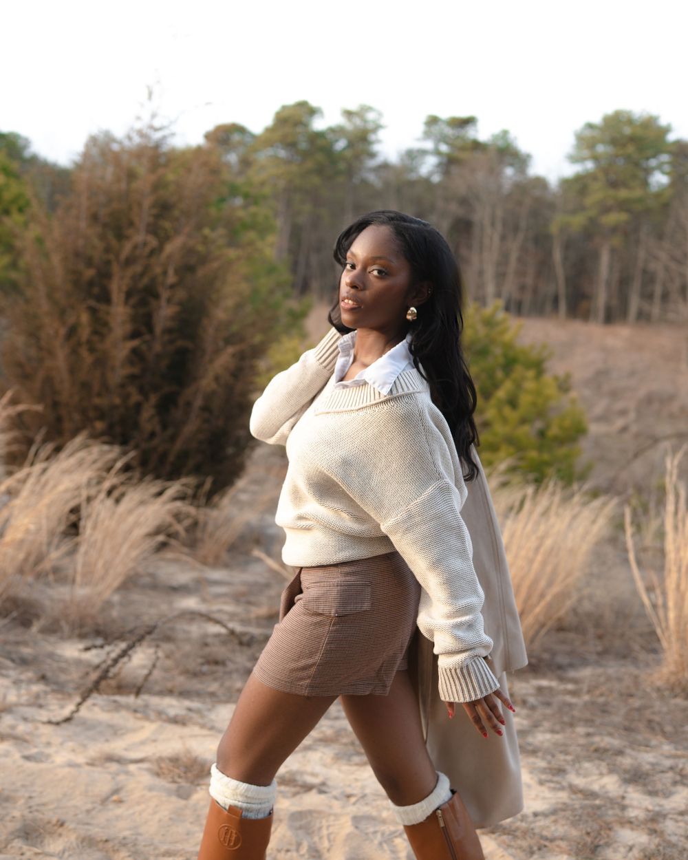 Stylish winter outfit featuring a cream knit sweater and tall brown boots amid dried grass and pine trees in natural scenery.