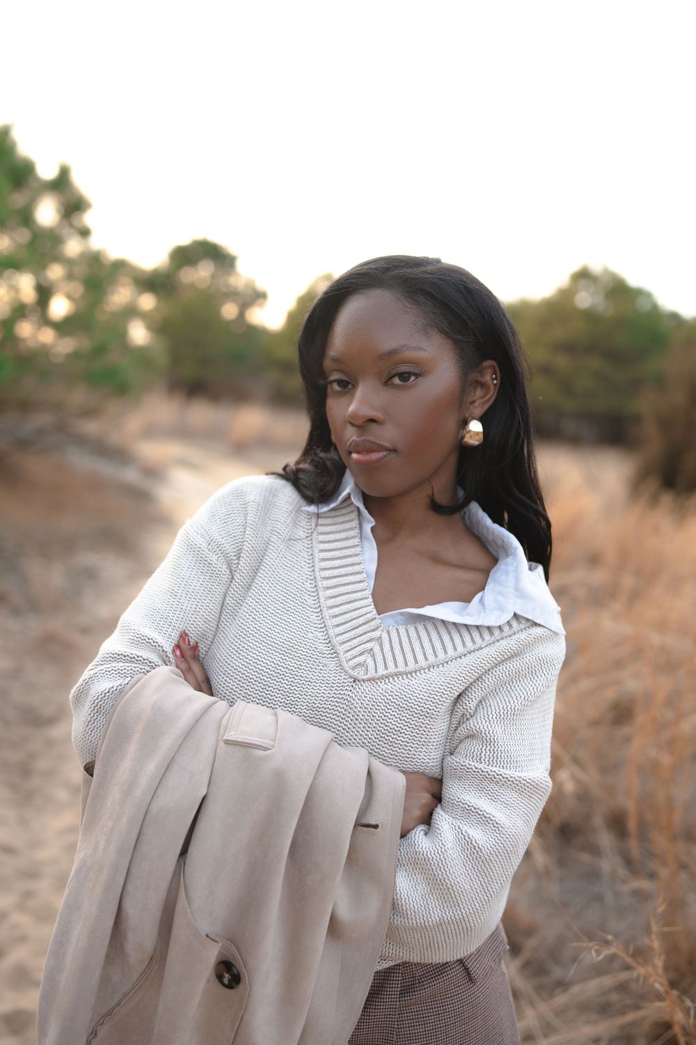 Fashionable woman in white knit sweater and pearl earrings holding beige coat while posing outdoors at sunset.