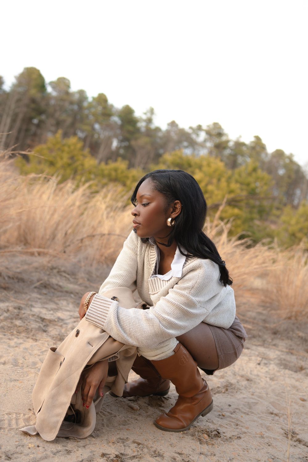 A person in a cream sweater and brown boots crouches in a scenic area with tall grass and pine trees in the background.