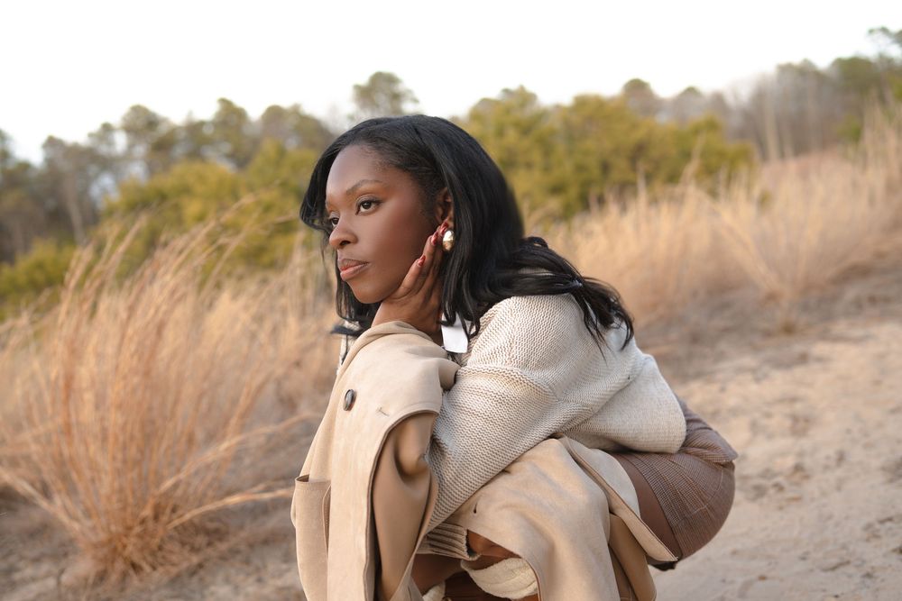 Person in a beige coat poses outdoors among tall dried grass during golden hour.