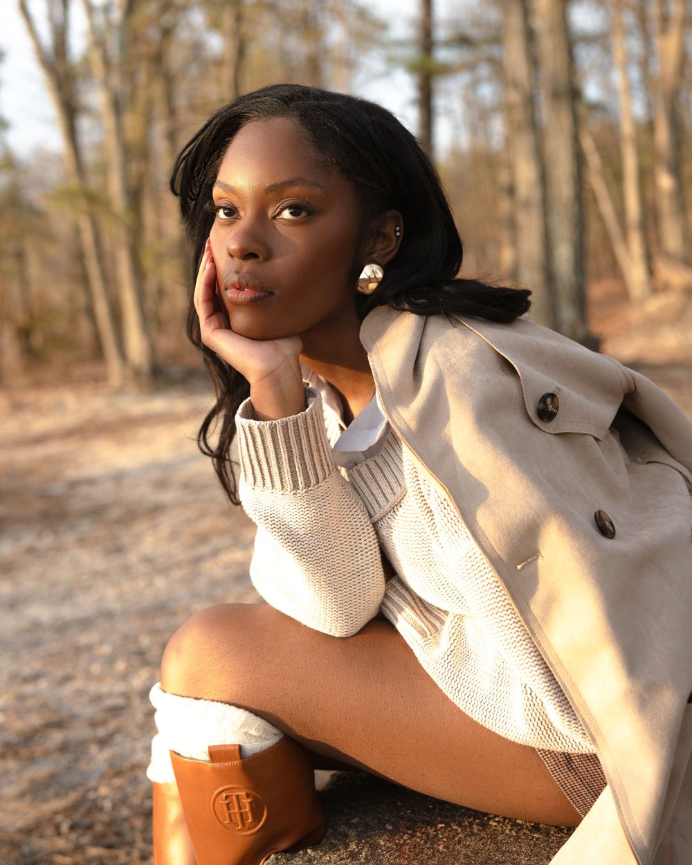 Person wearing cream sweater, beige jacket and brown boots poses thoughtfully in a sunlit autumn forest setting.