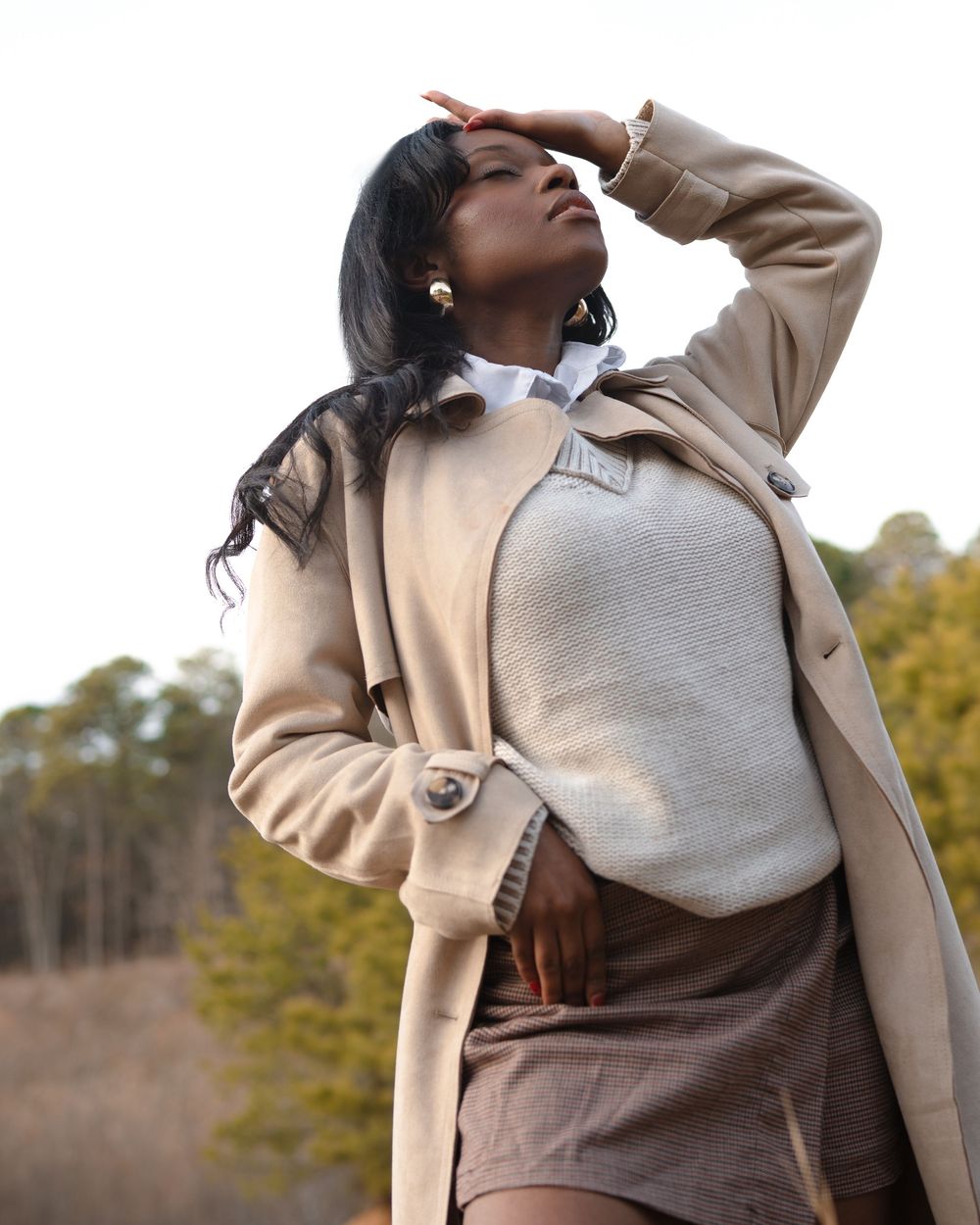 A person in a beige coat and gray hoodie poses outdoors with head tilted back in a stylish autumn outfit.