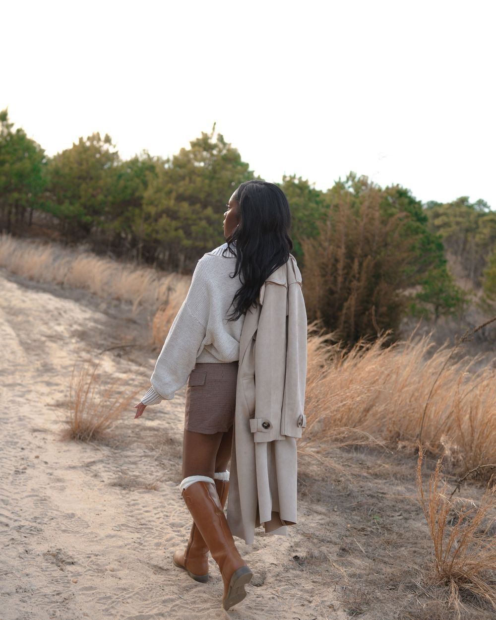 Woman in beige trench coat and brown boots walking on sandy path through tall grass and pine trees.