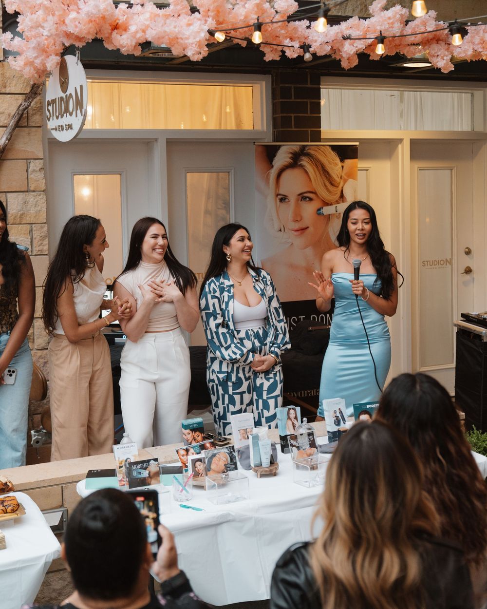 A group celebrates at an outdoor party with pink cherry blossoms overhead and an elegant table setting.