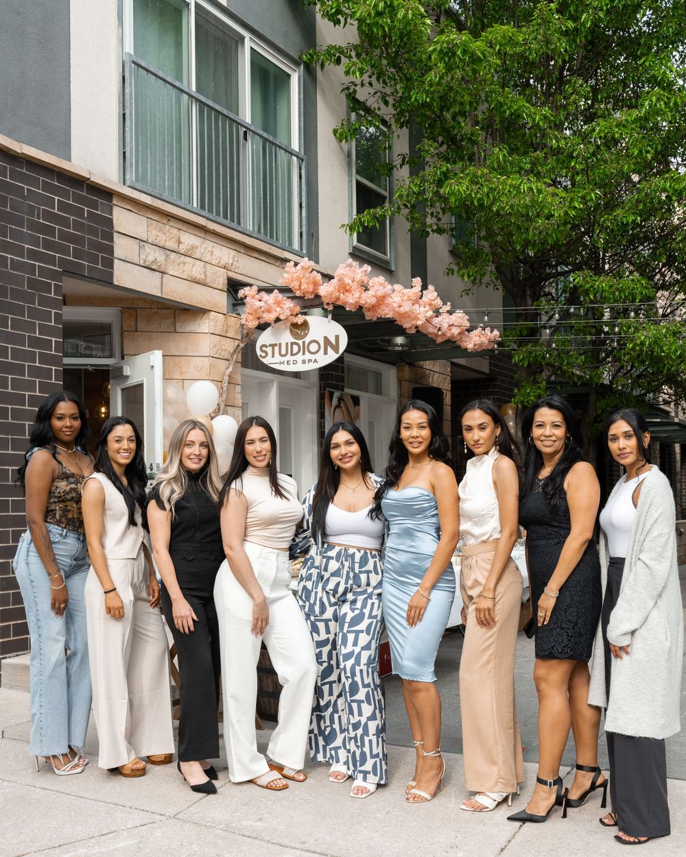 Group of women in stylish outfits standing outdoors at an elegant storefront decorated with pink floral arrangements.