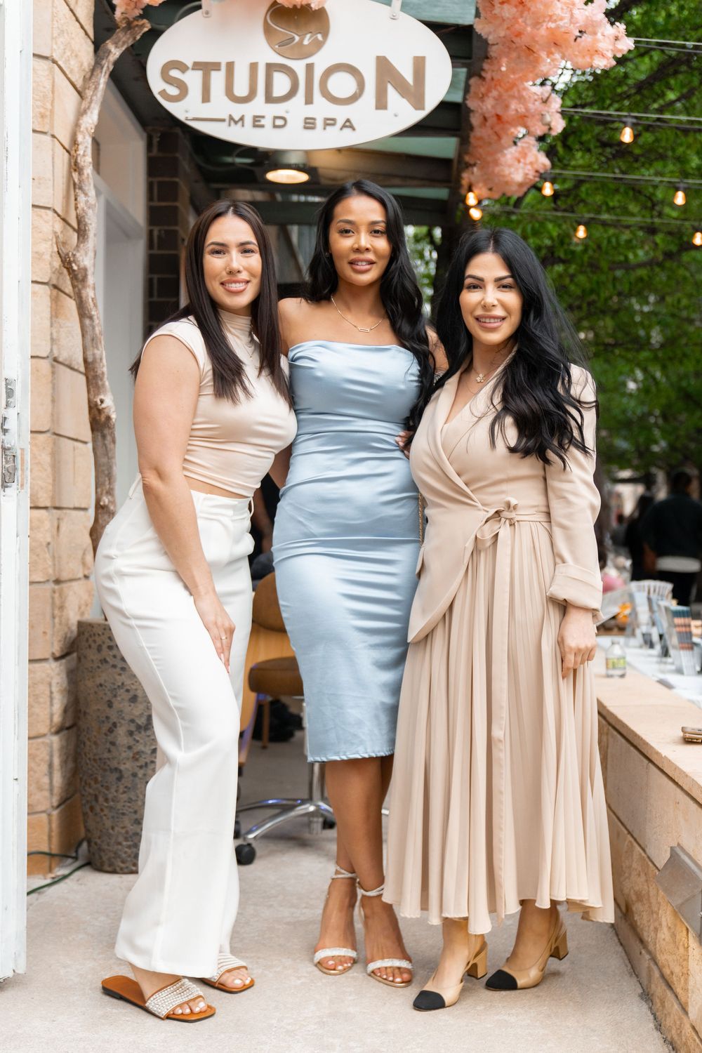 Three friends pose together outside Studio N med spa wearing elegant dresses in white, blue and beige tones.