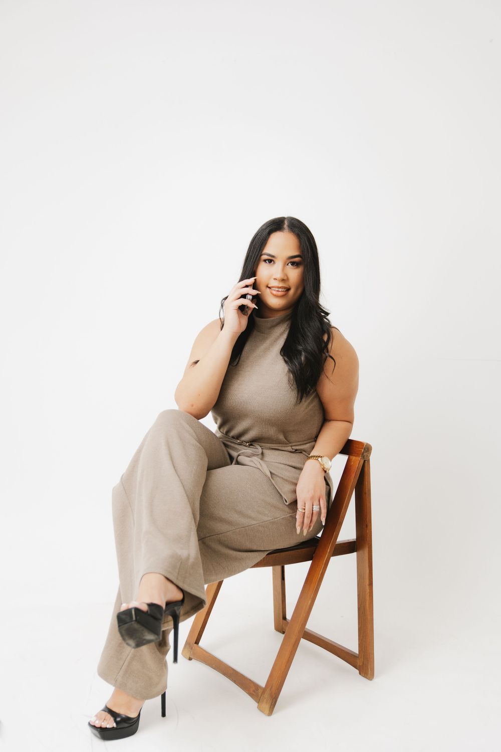 Woman in beige outfit talking on phone while seated in wooden chair.