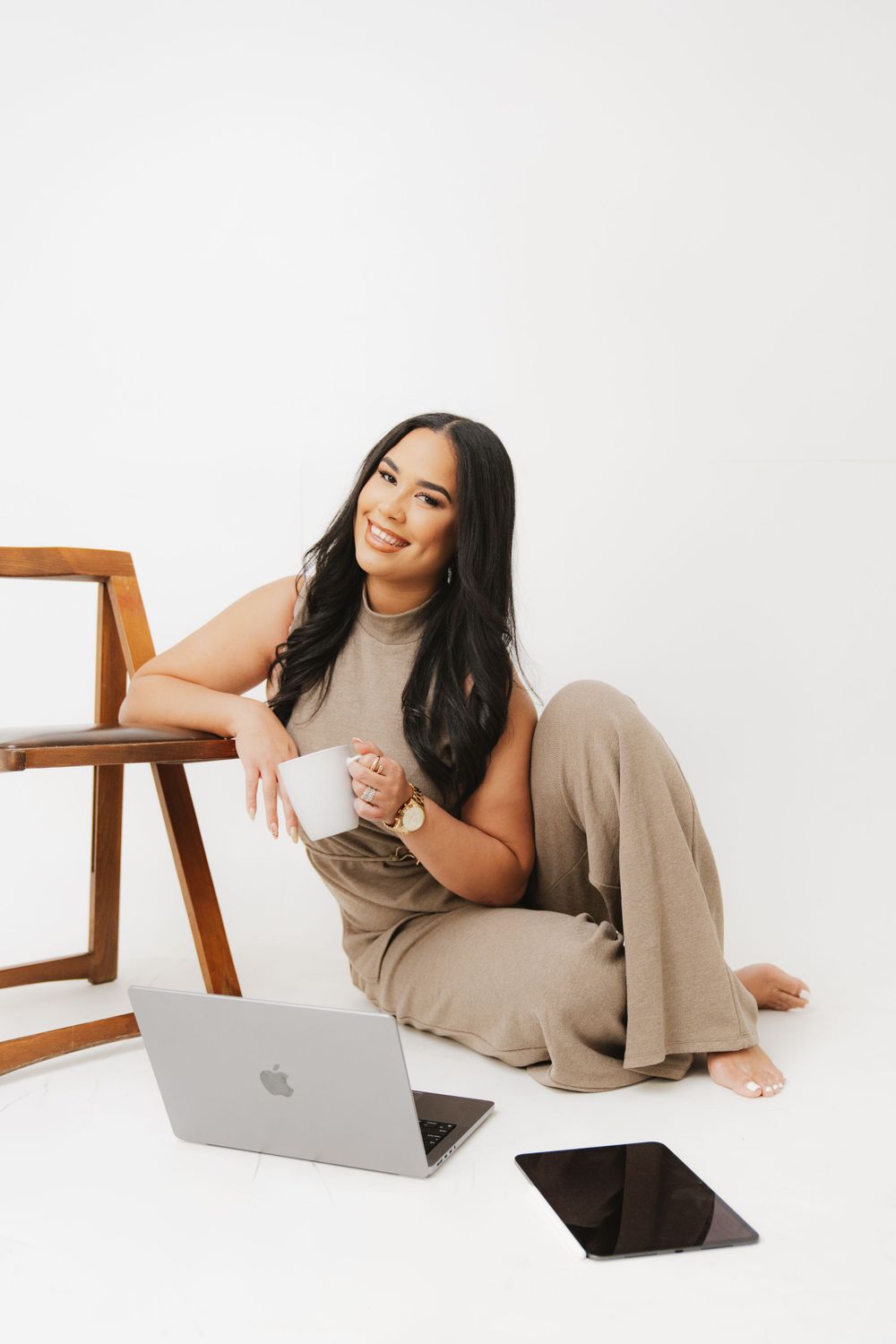 Woman in casual attire working on laptop while sitting on floor next to wooden chair.
