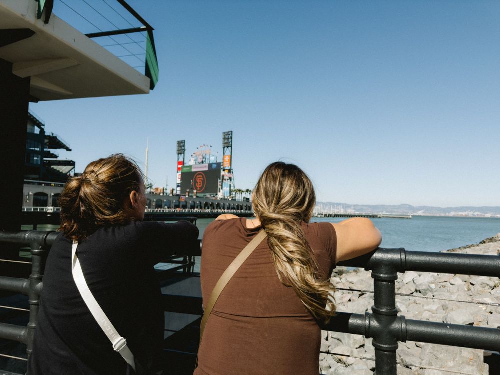 Two women lean on a railing overlooking a baseball stadium by the bay on a clear sunny day.