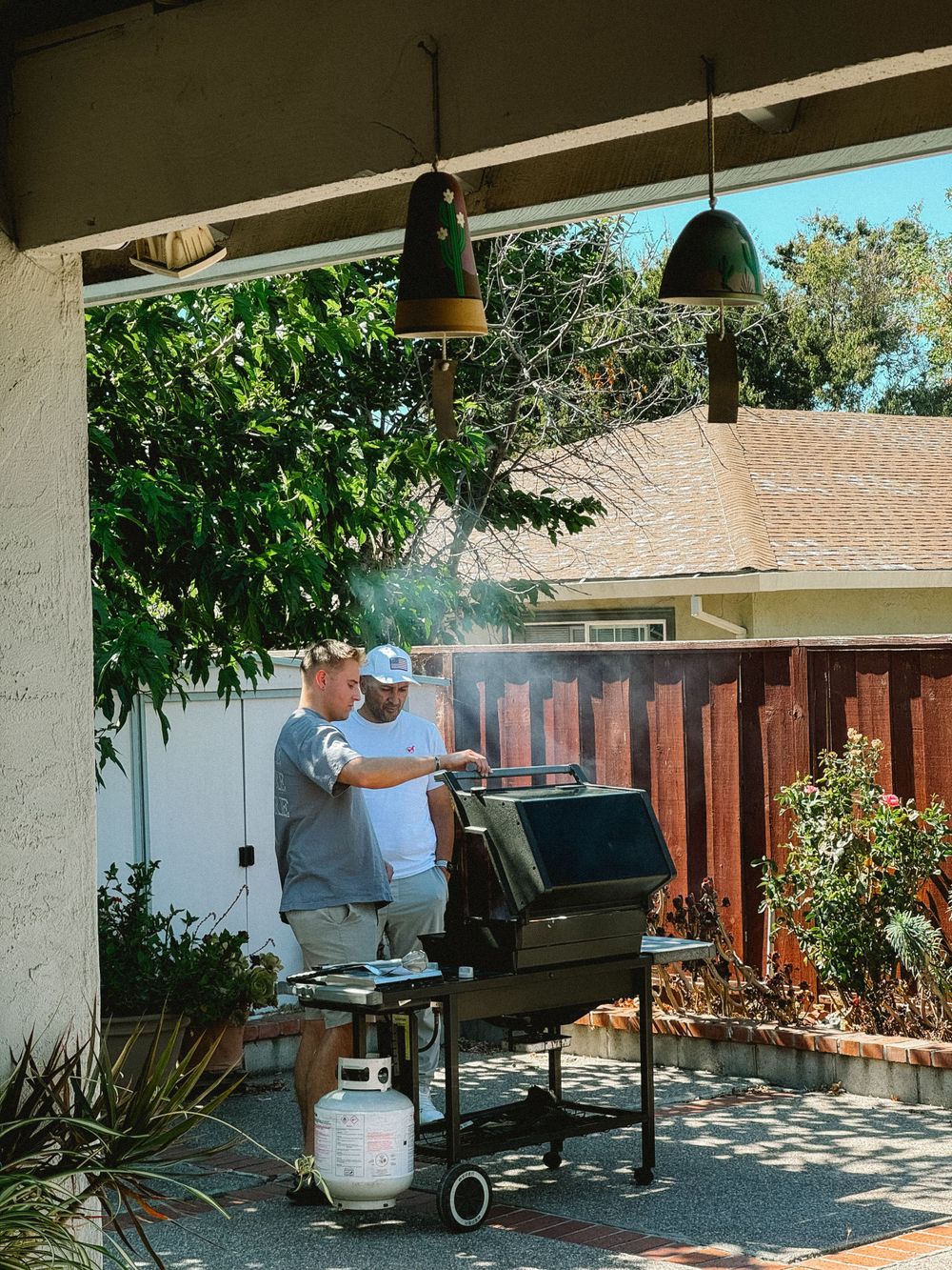 Two men grilling outdoors on a large smoker BBQ grill under a covered patio.