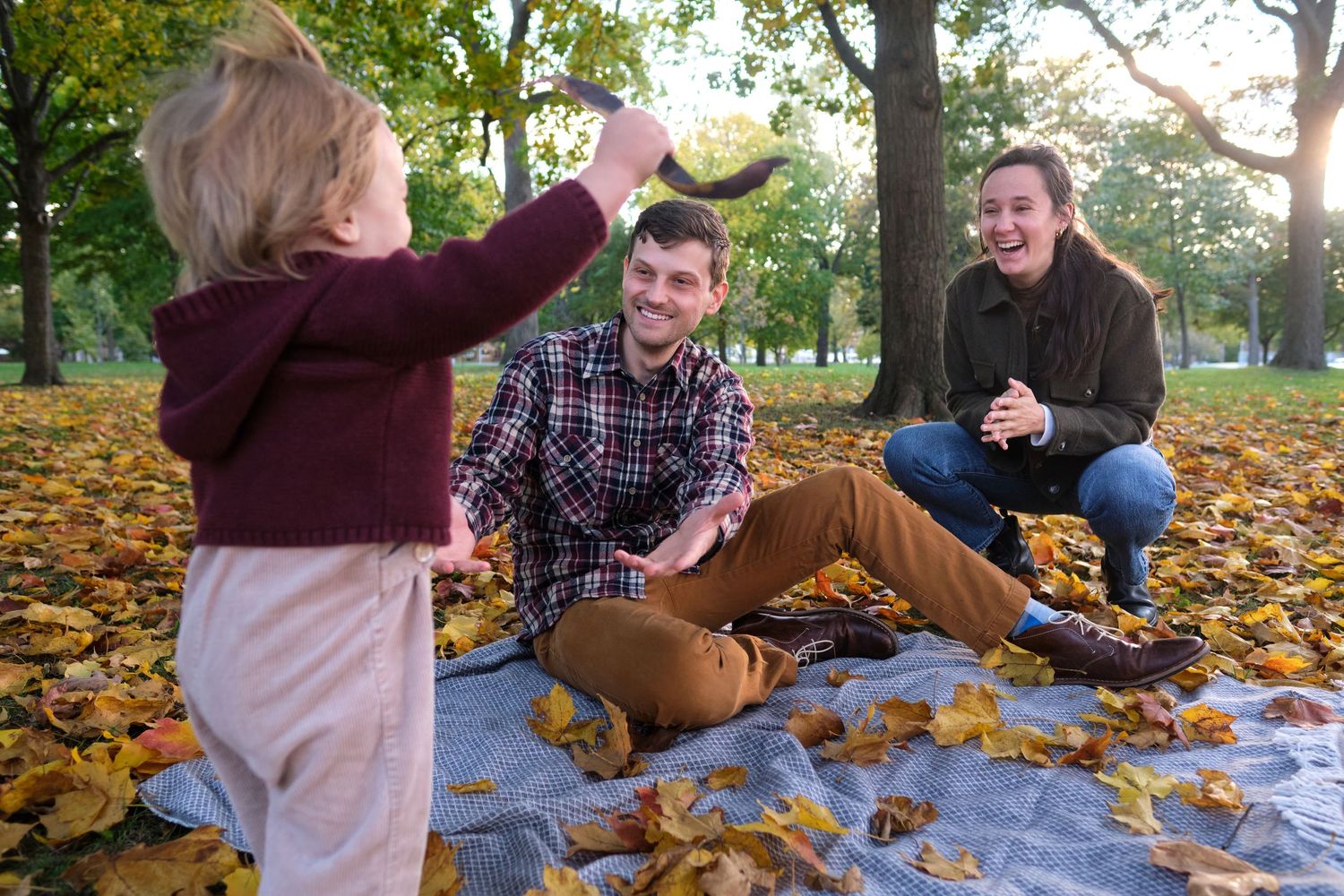 A family photo of a man, woman and their child sitting on a blanket surrounded by fall colors.
