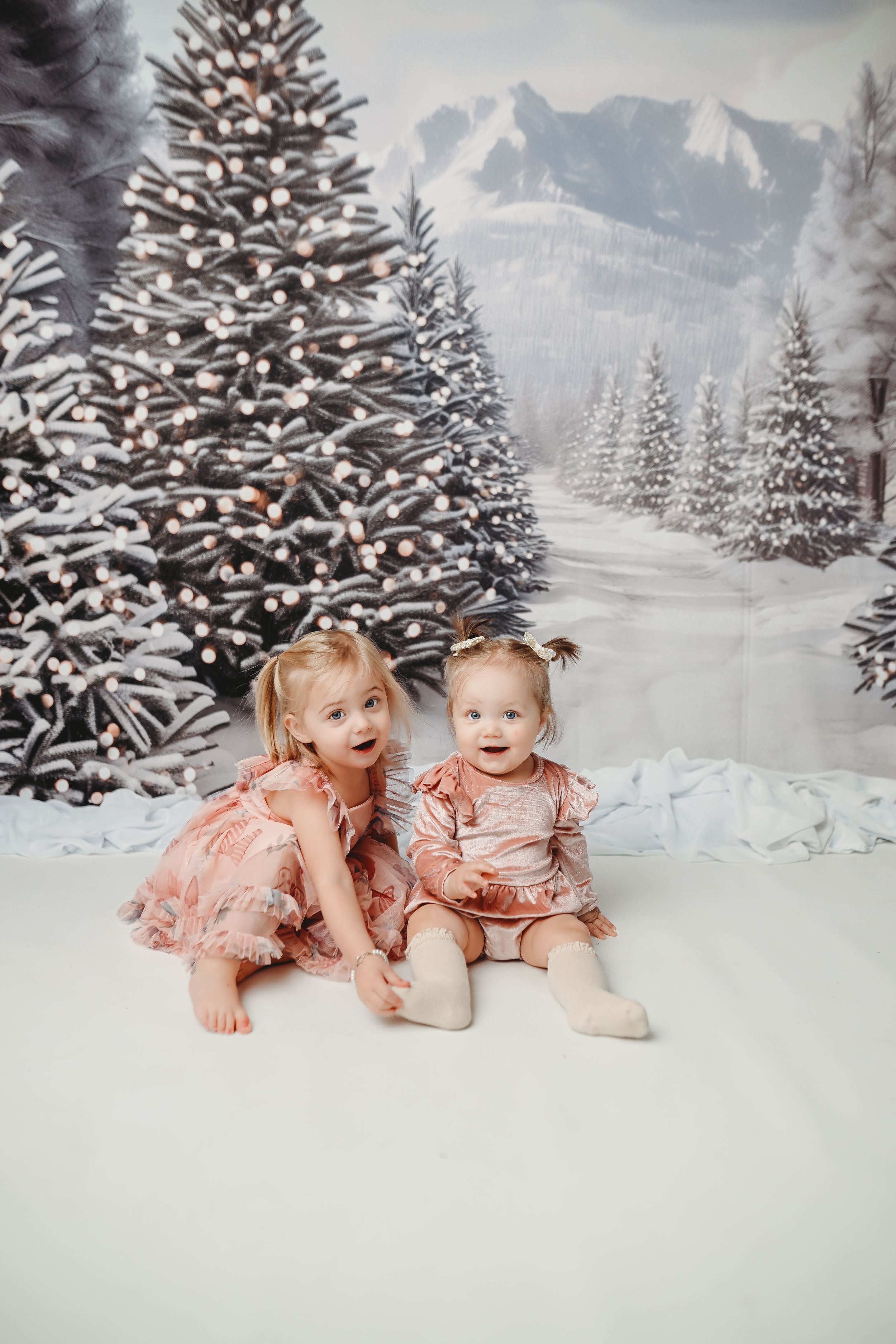 Two children in pink dresses sitting against a snowy winter wonderland backdrop with decorated trees.