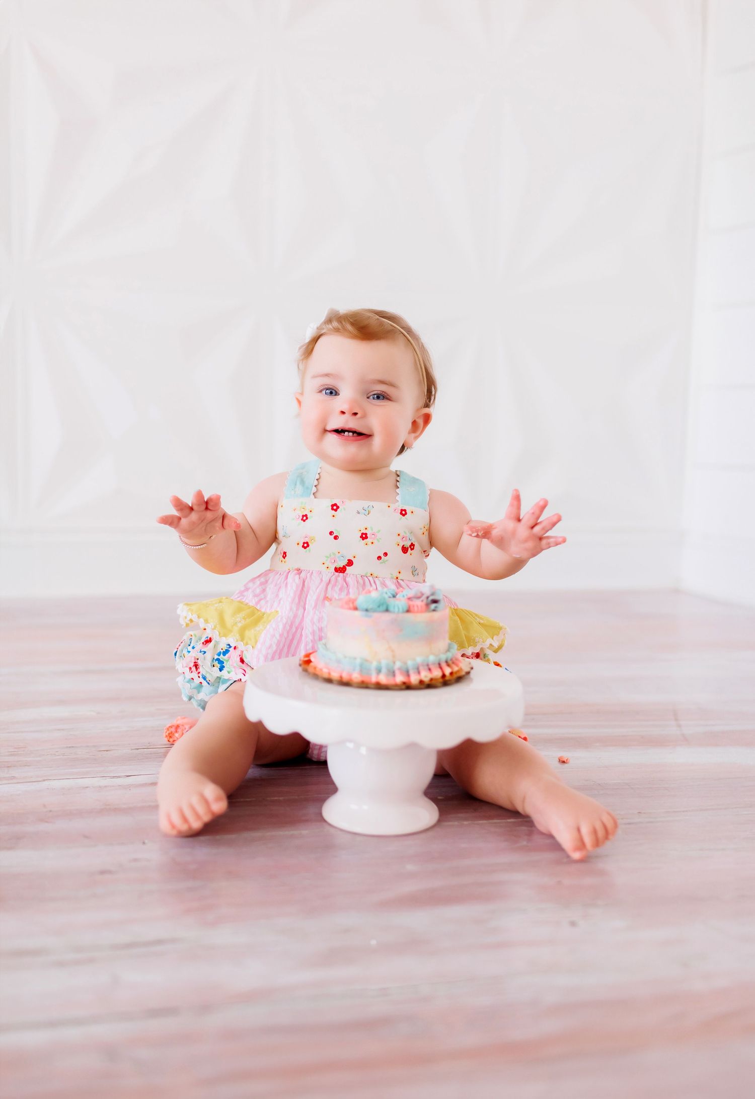 Happy baby in floral dress sitting next to small cake on wooden floor.