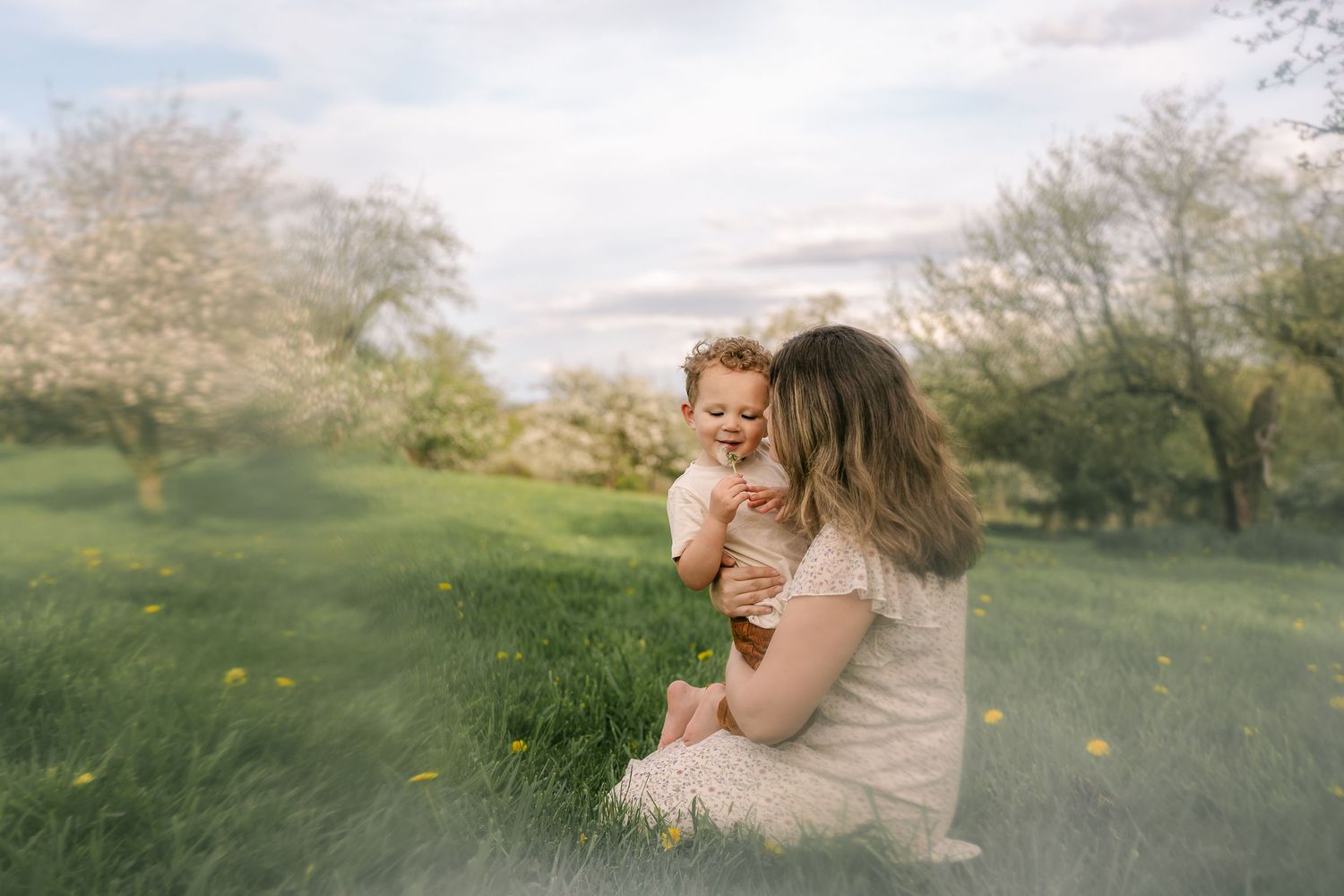Mother and child share tender moment in spring meadow with blooming trees.