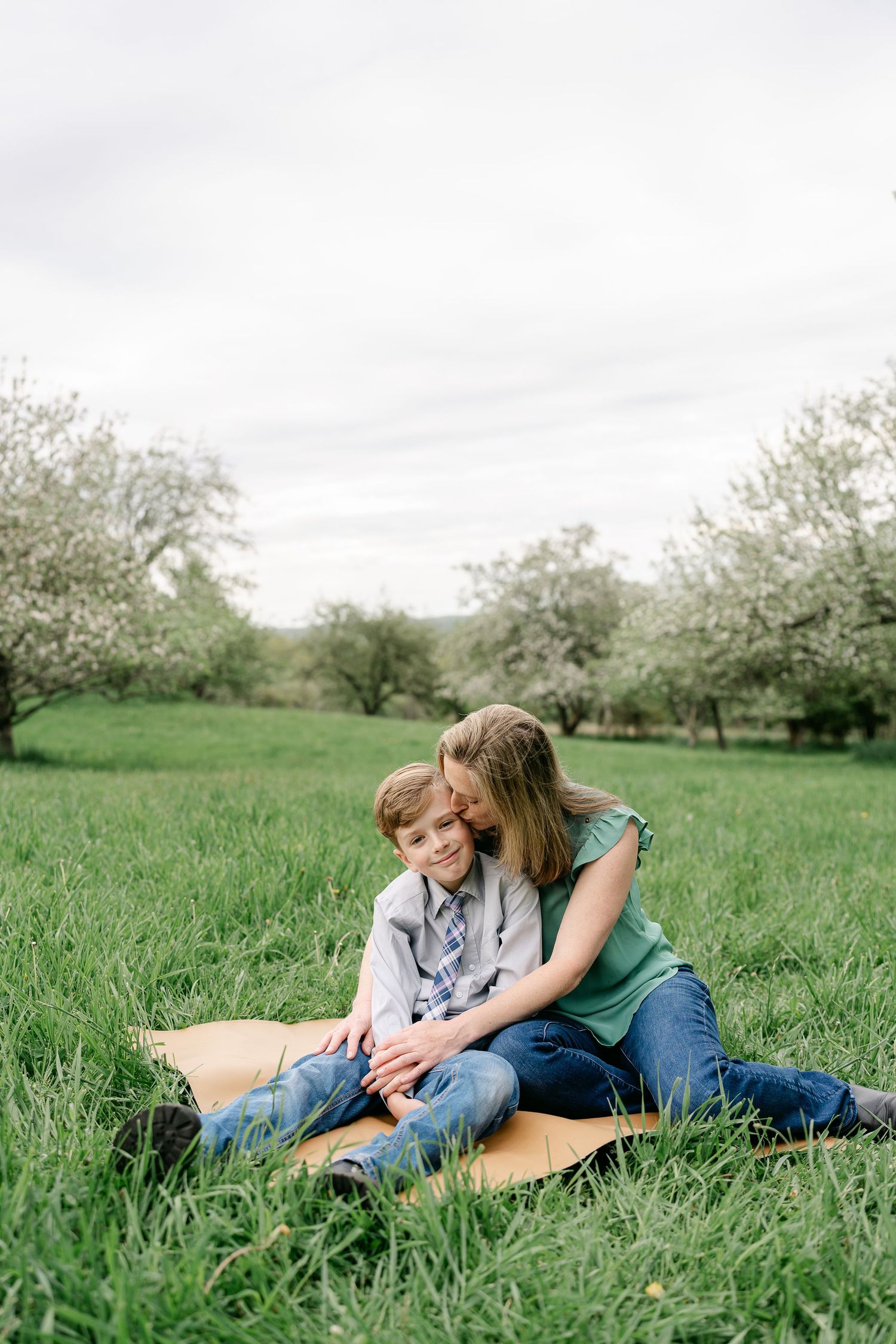 A tender moment shared on a picnic blanket in a blooming apple orchard with tall grass and overcast sky.