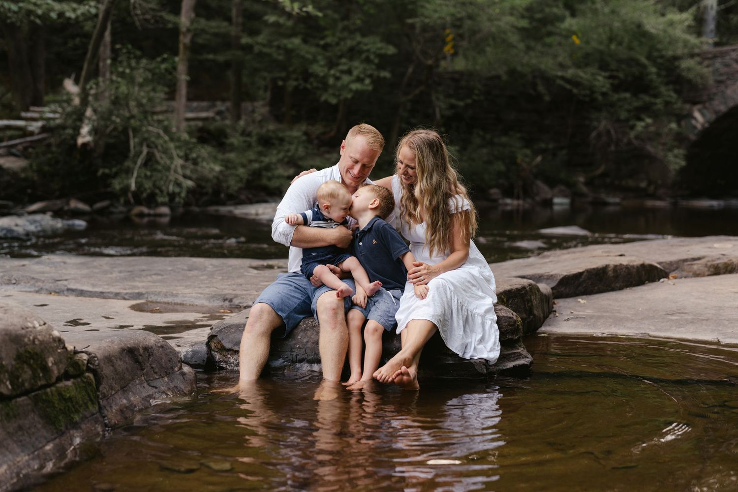 Family sitting together on rocks by a stream in a wooded setting enjoying a peaceful moment.