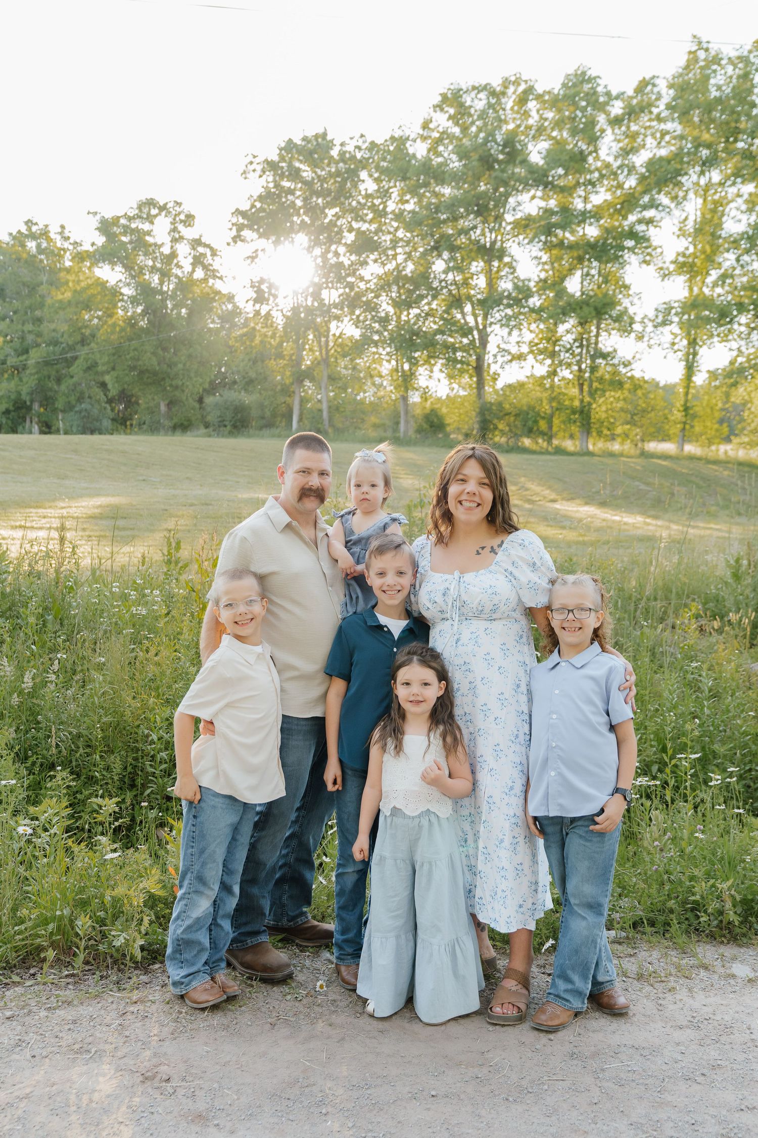 Family of seven posing outdoors at golden hour in a lush green field, parents with five young children smiling.