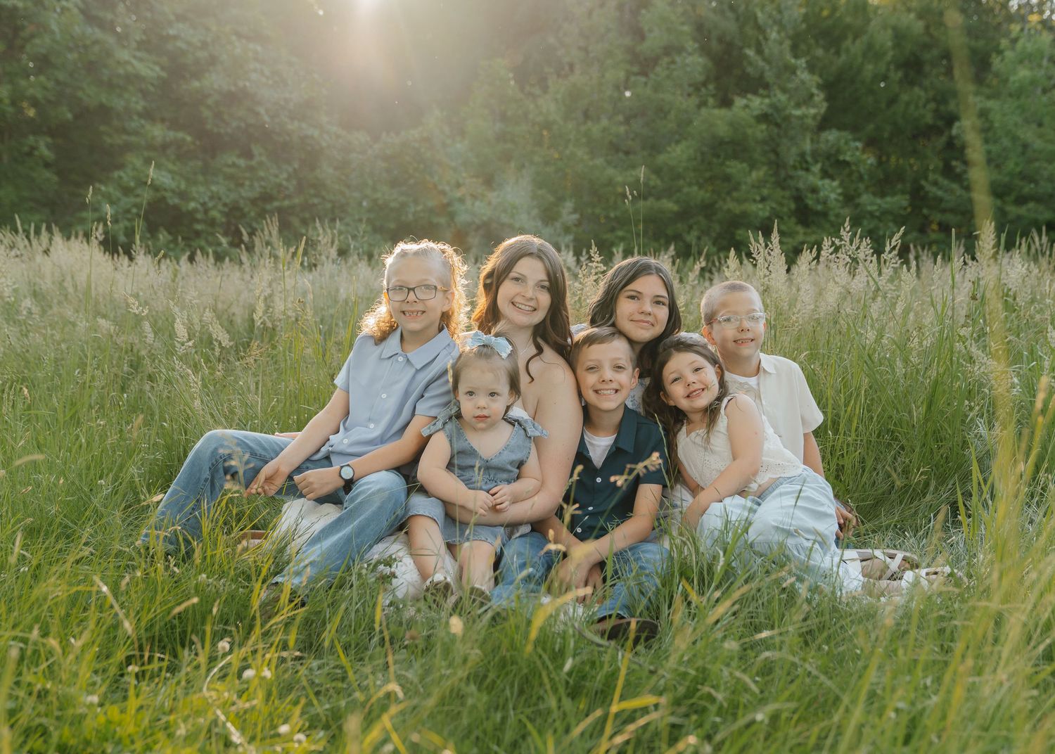 Multi-generational family portrait of seven people sitting together in a sunlit meadow with tall green grass.