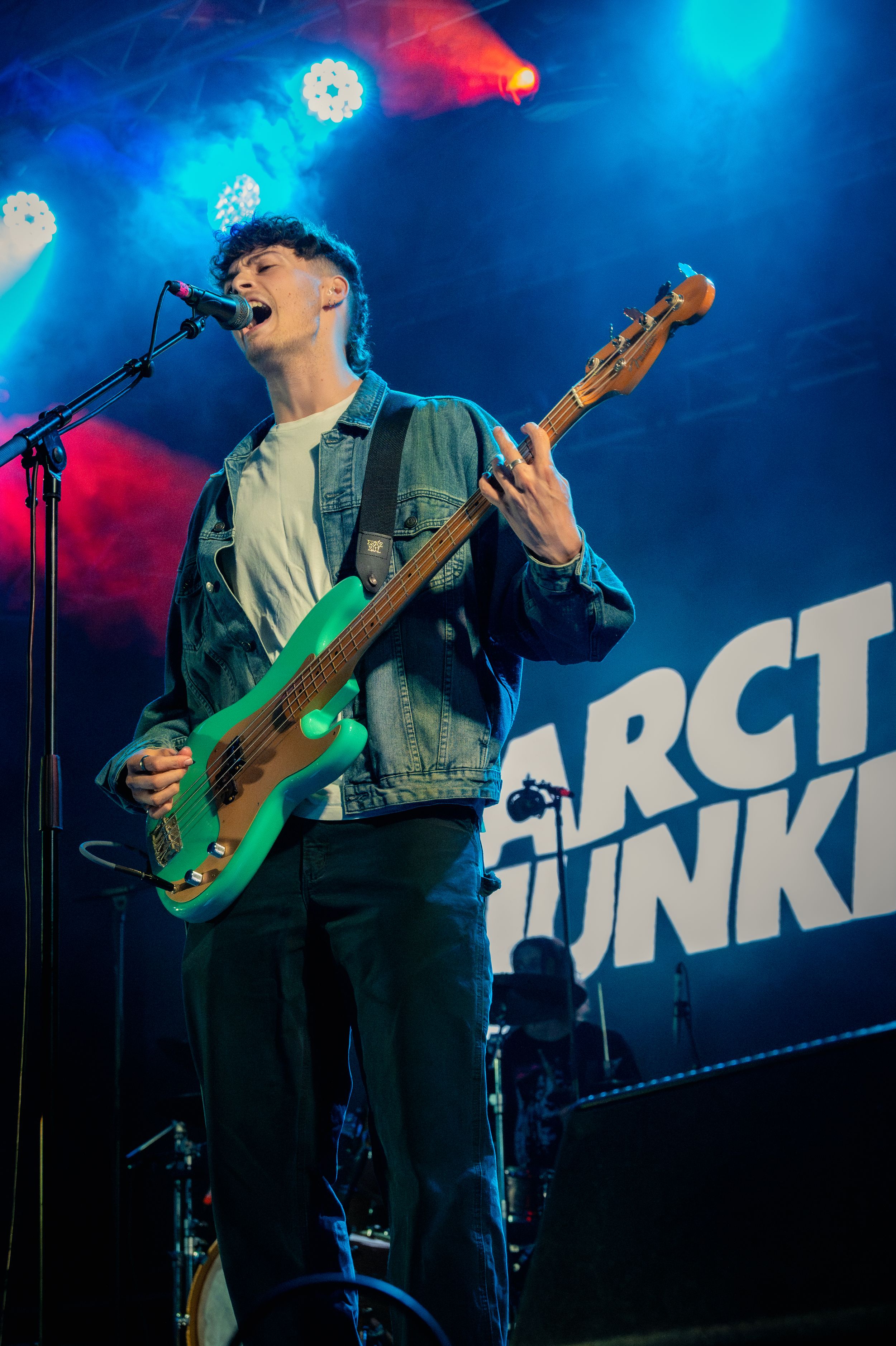 Bass guitarist performing on stage with blue lighting and Arctic Junks backdrop.