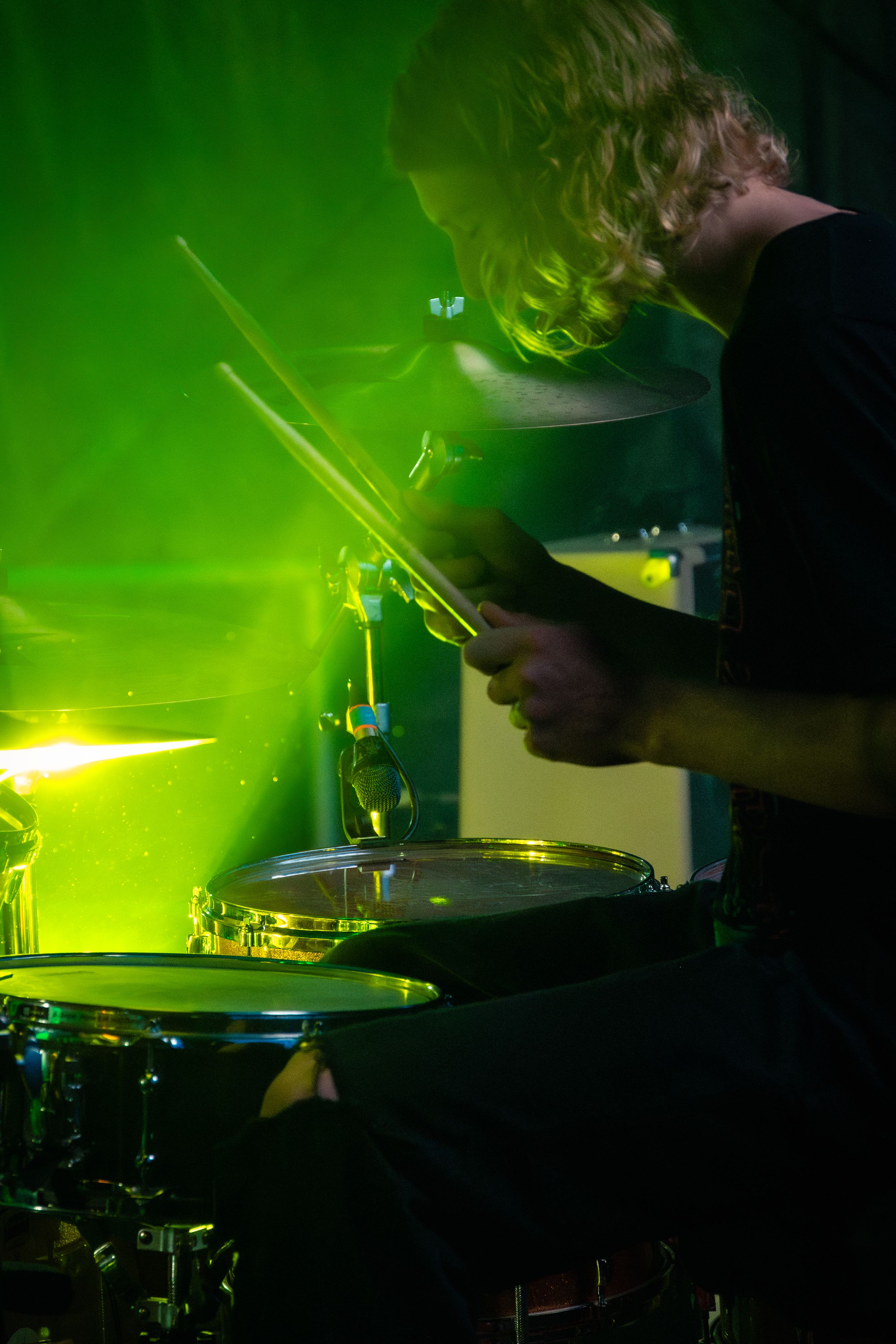 Drummer performing on stage illuminated by vibrant green lighting effects.