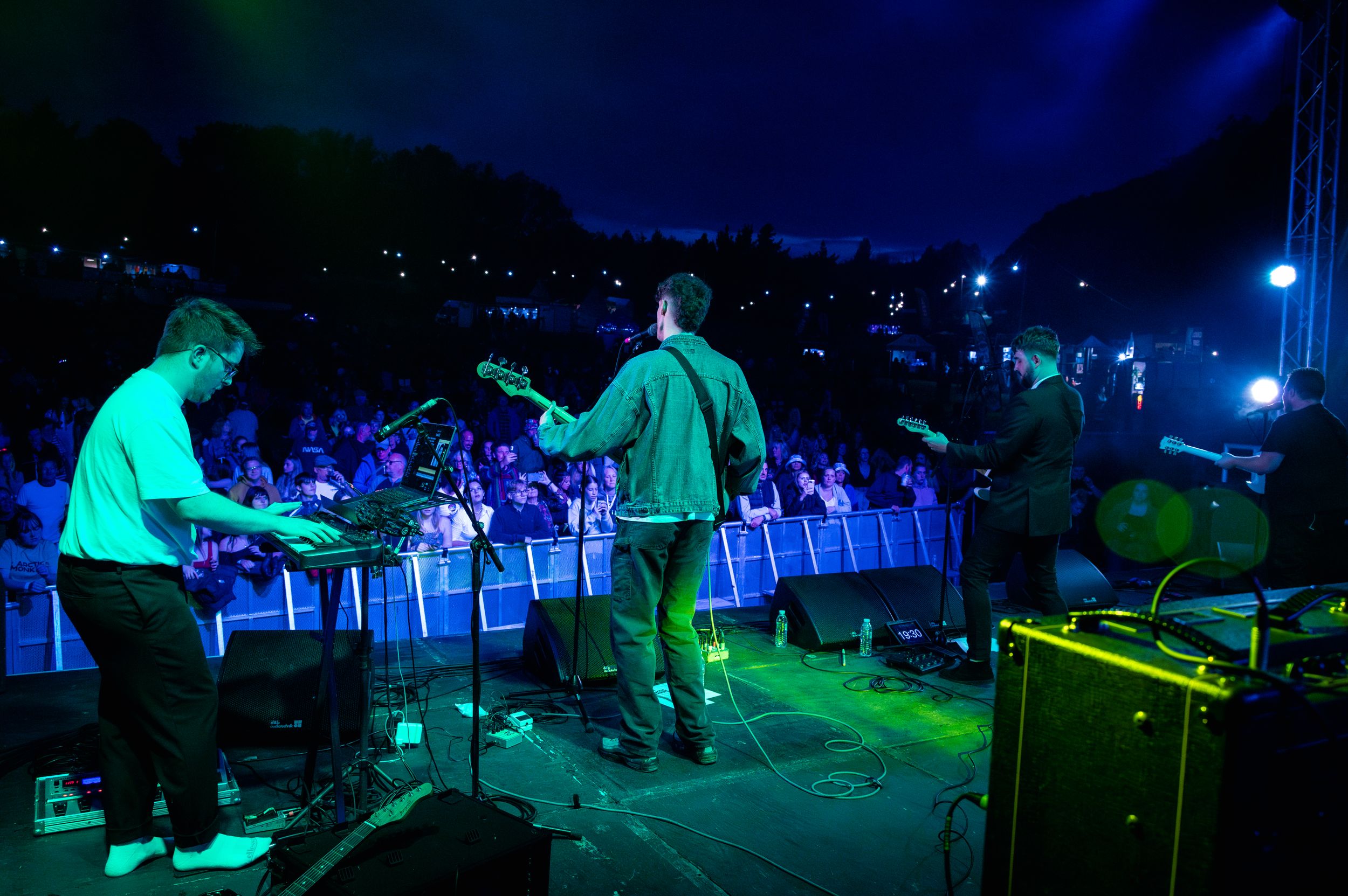 Band performing on outdoor stage with blue lighting and crowd barriers visible.