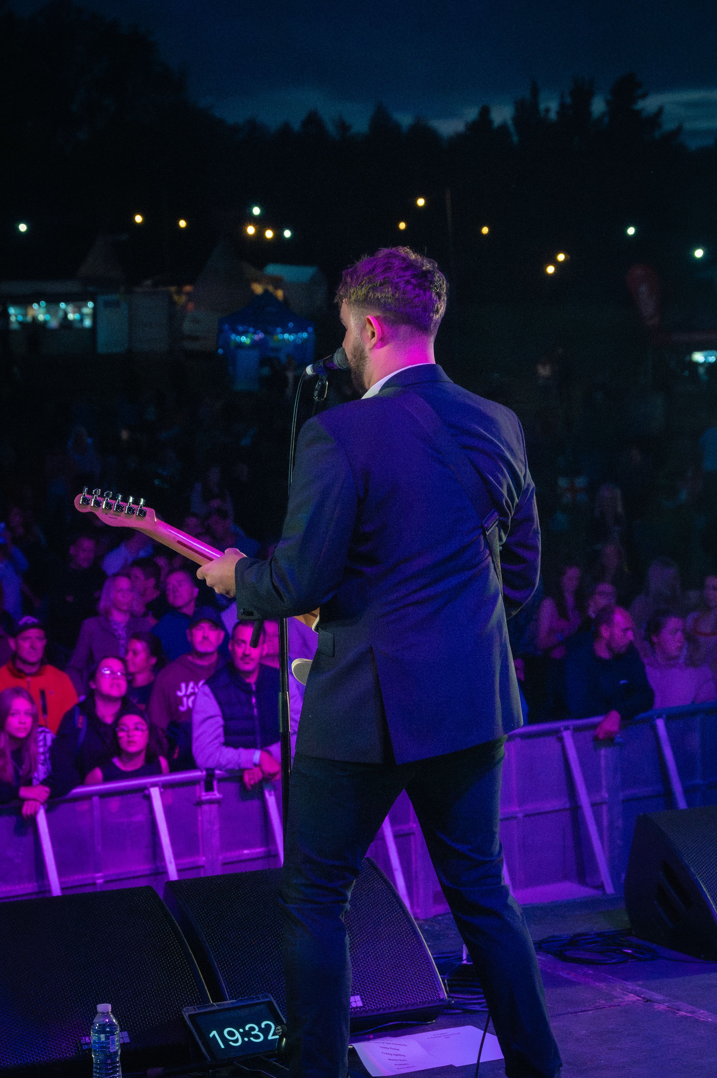 Performer in suit jacket on stage with colorful lighting and crowd in background.