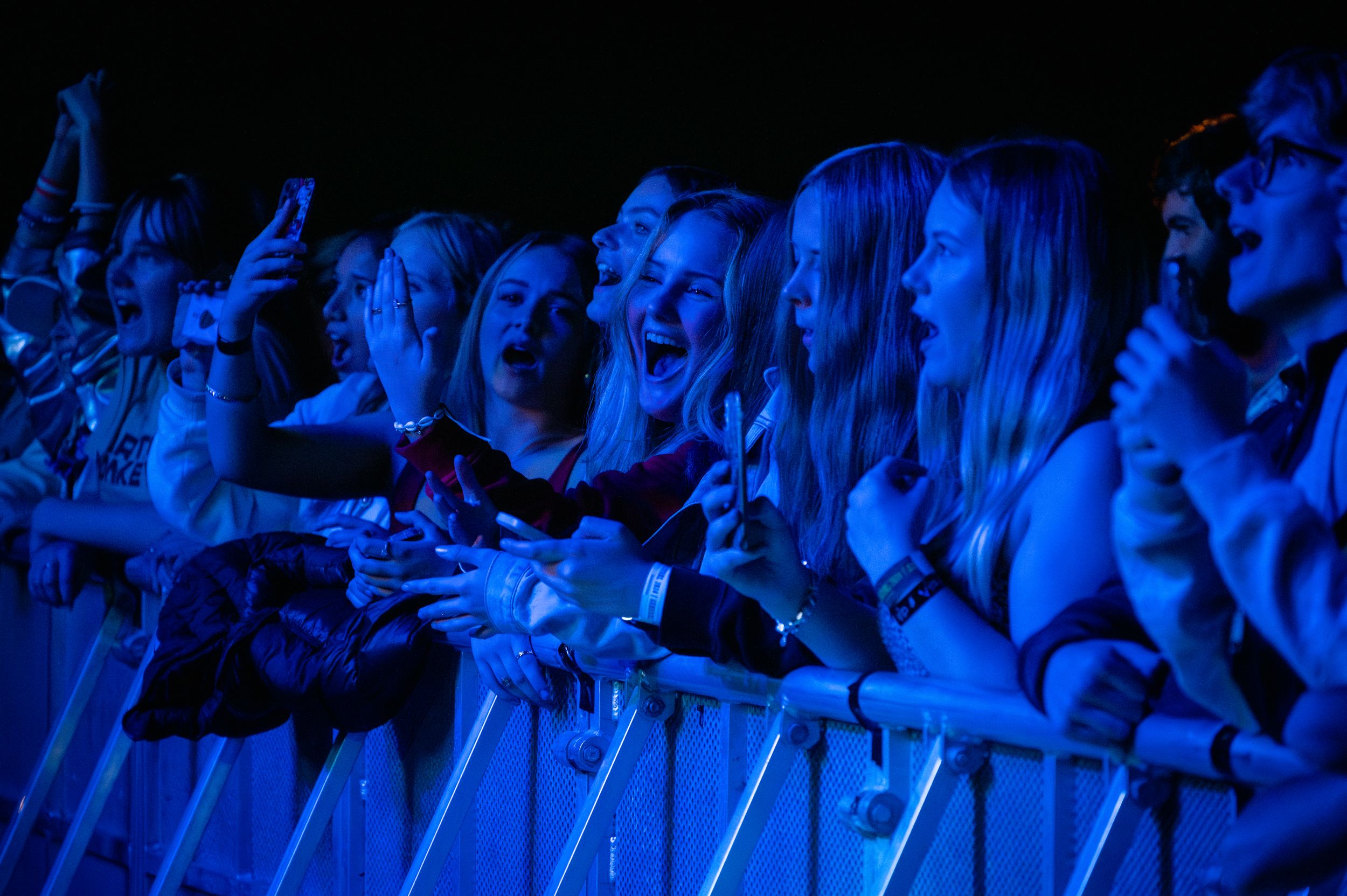 Concert crowd illuminated in blue light cheers enthusiastically at front row barrier.