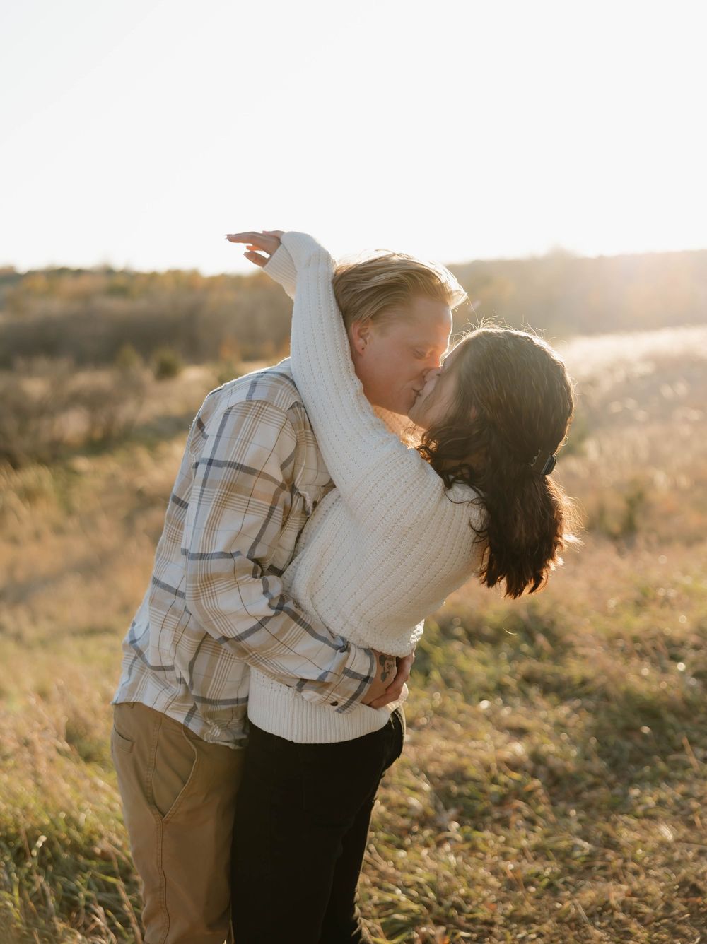 Two people embrace in a golden sunlit field during sunset while wearing casual clothing.
