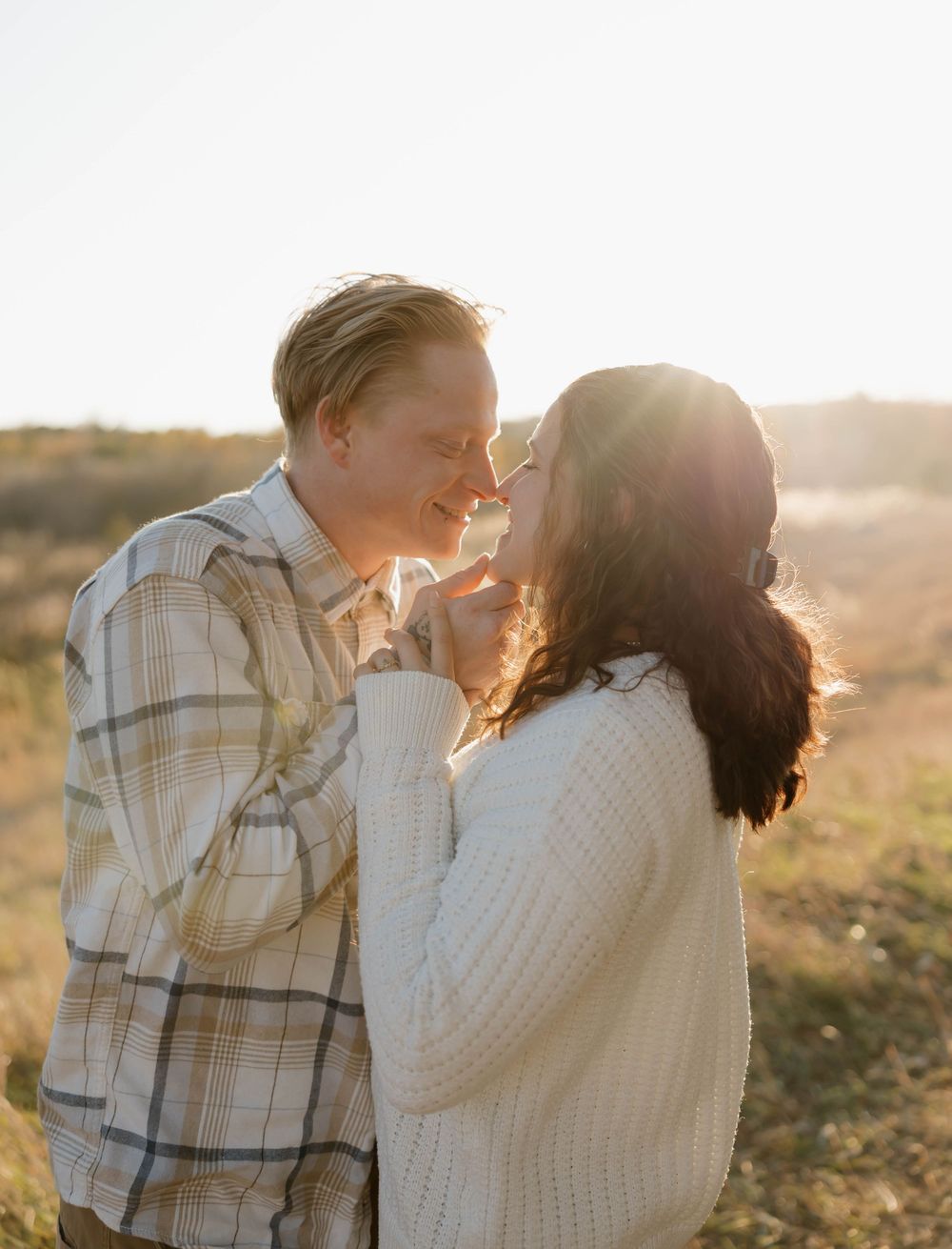 A couple shares an intimate moment at sunset in a field wearing plaid and knit sweaters.
