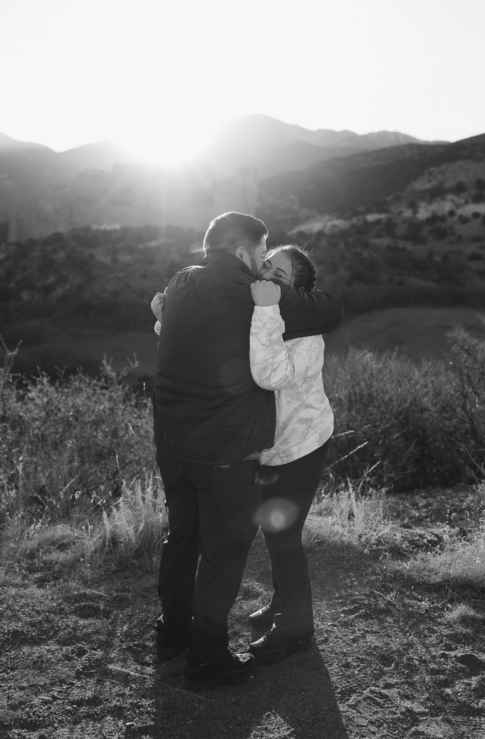 Two people embrace in a sunlit field with mountains and golden grass creating a romantic silhouette.