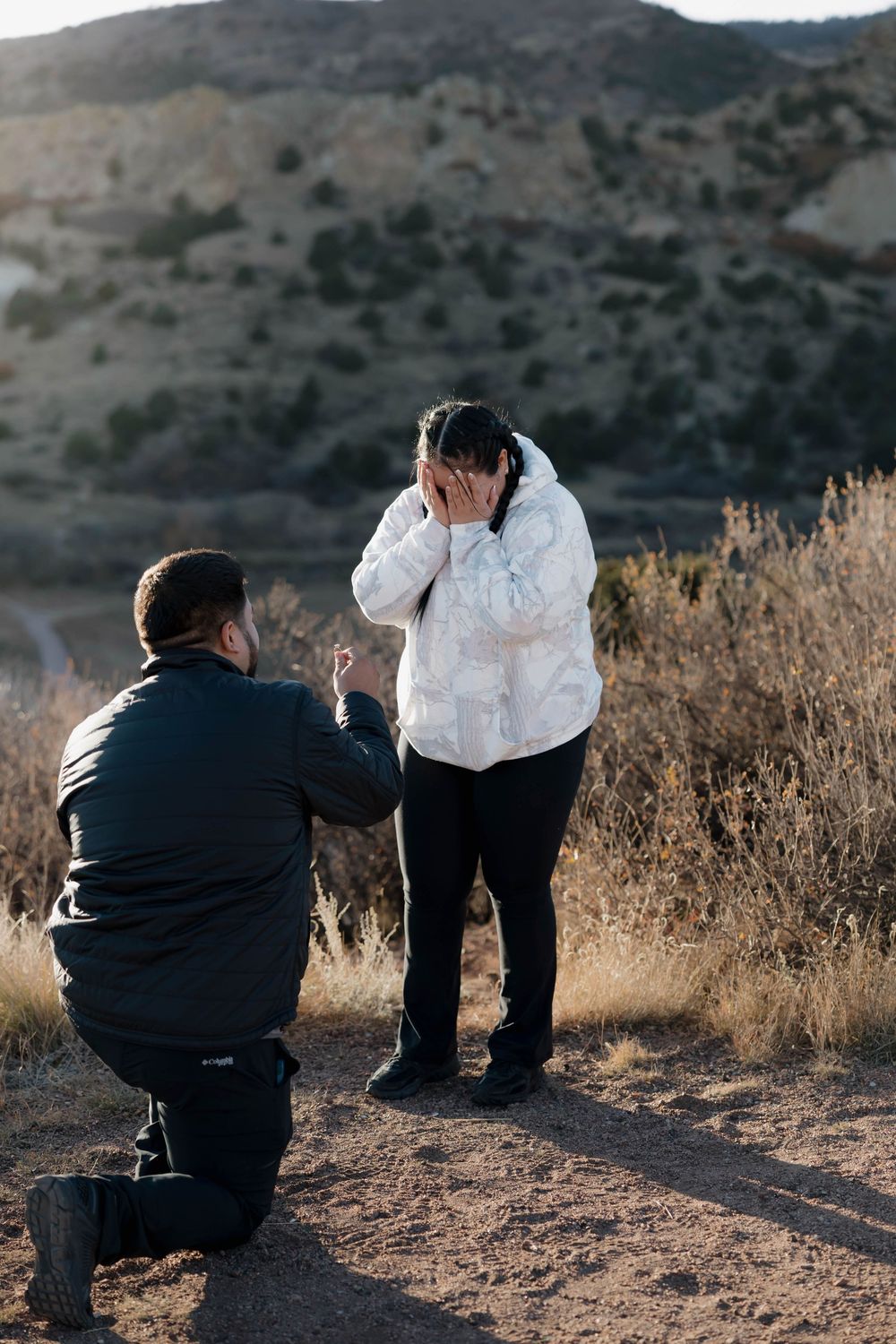 A couple in casual attire shares a tender moment outdoors against a backdrop of dry brush and hills at sunset.