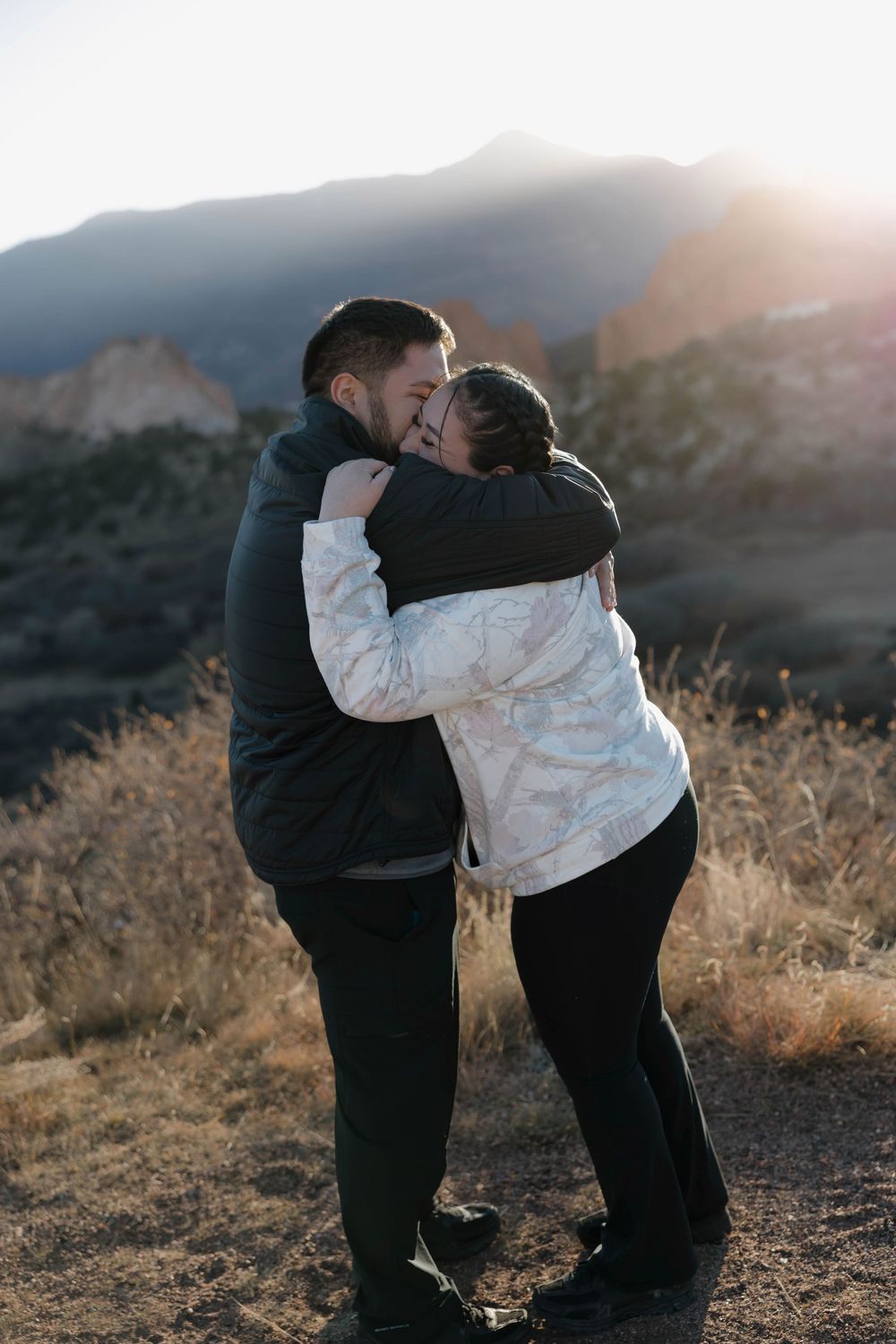 A couple embraces during sunset in a mountainous landscape with golden light and dry brush in the background.