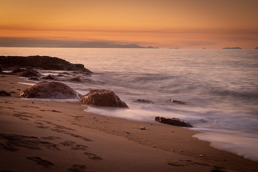 Waves crash against rocky outcrops on a sandy beach during a golden sunset over the ocean.