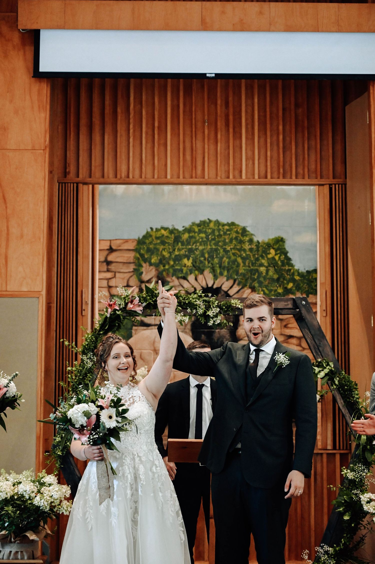 Newlyweds celebrate with raised arms at their wedding ceremony under a wooden archway decorated with flowers.