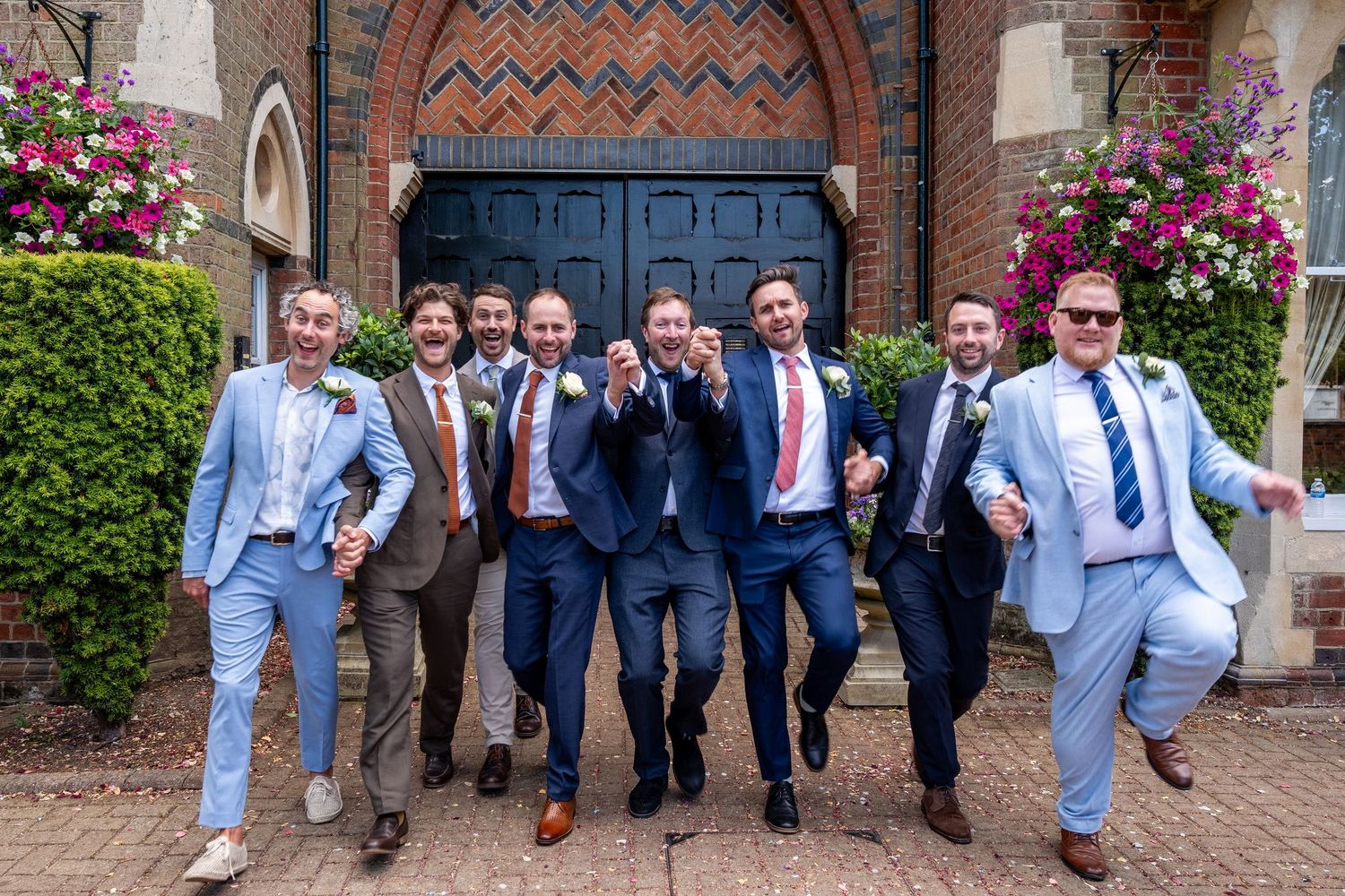Groomsmen in blue and navy suits celebrate joyfully outside a brick church with floral decorations.