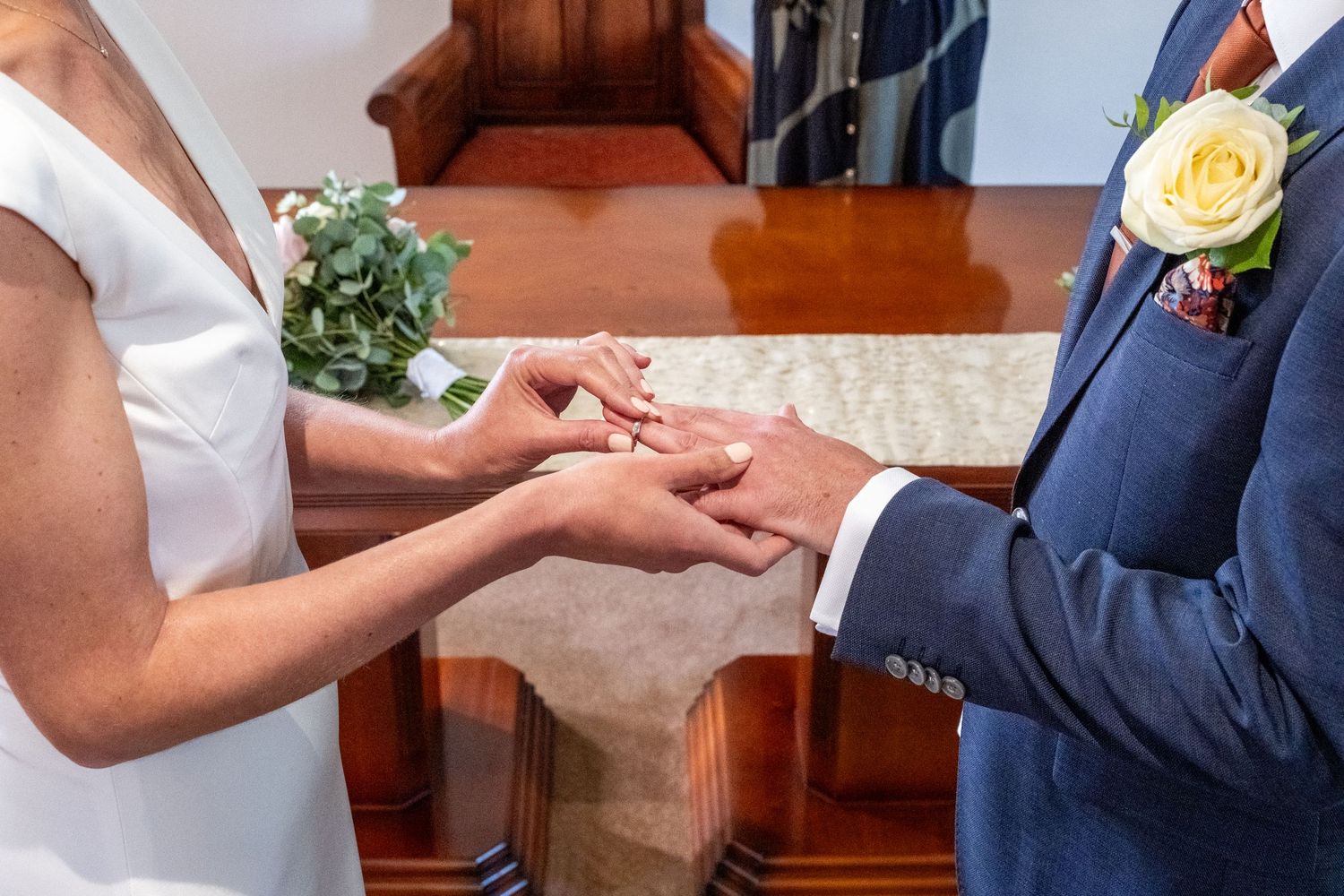Bride placing a wedding ring on groom's finger during ceremony, with bridal bouquet and blue suit visible.