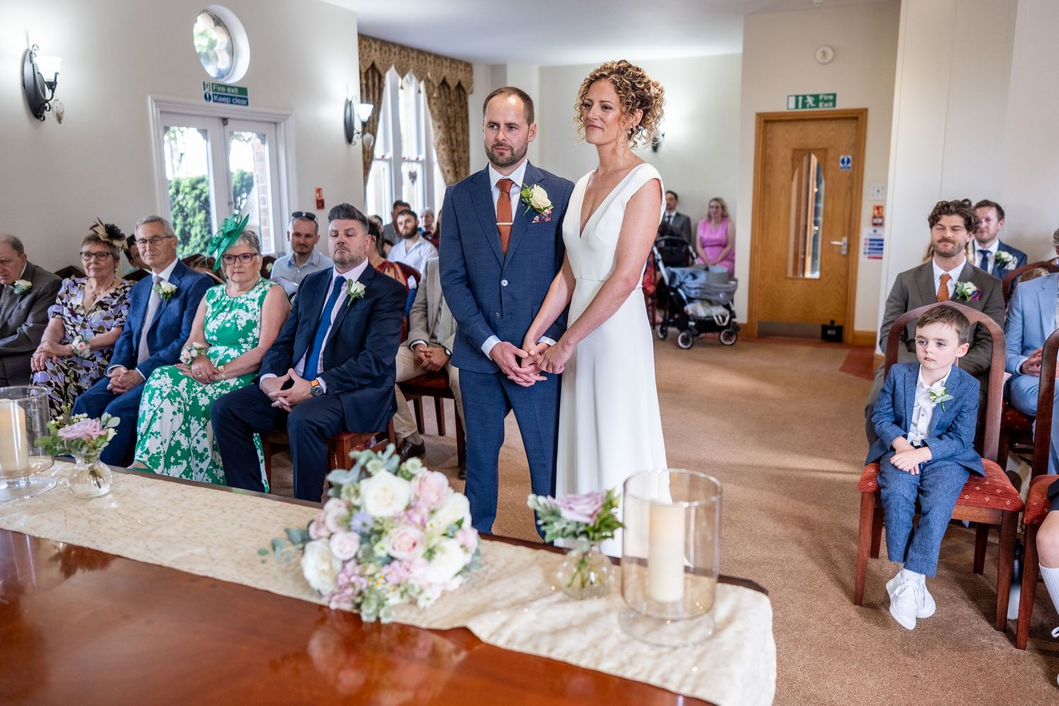 Bride and groom stand at the altar during an intimate indoor wedding ceremony with seated guests in a bright, decorated room.