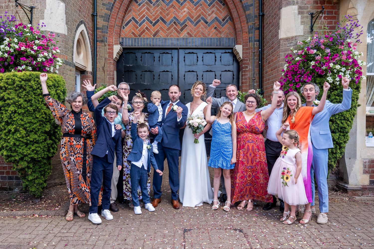 A joyful wedding group photo with bride and groom surrounded by family and friends outside a brick building with floral decor.