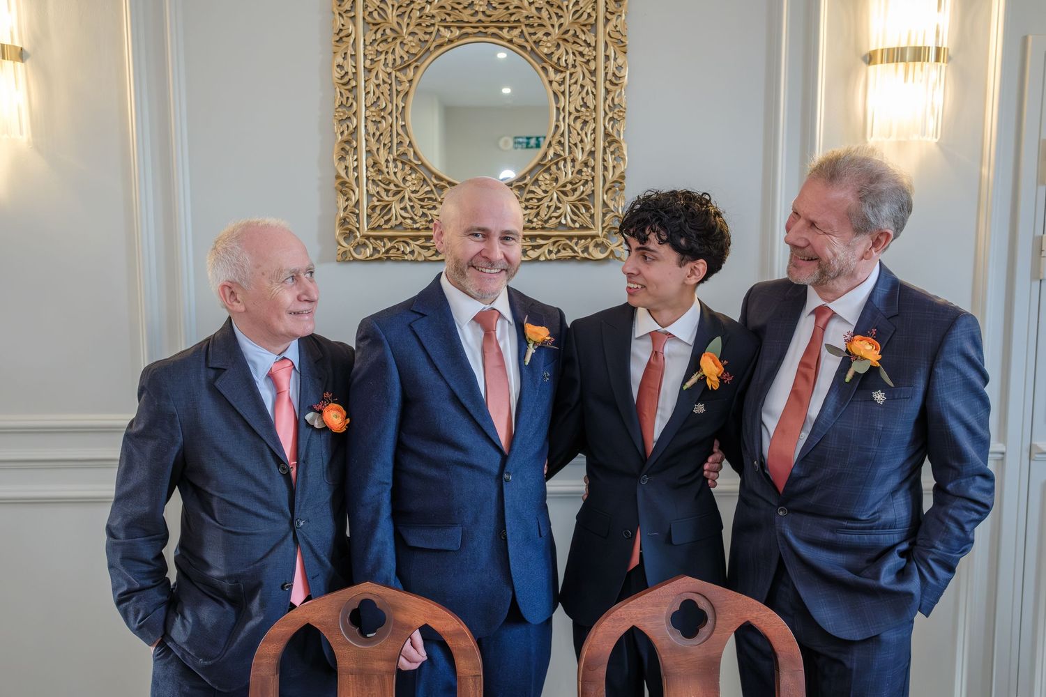 Four men in navy suits with orange boutonnieres smiling together at a formal wedding celebration indoors.