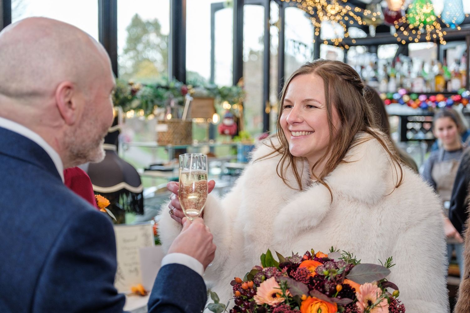 Bride in white fur wrap toasts champagne with groom while holding autumn bouquet at elegant indoor wedding venue.