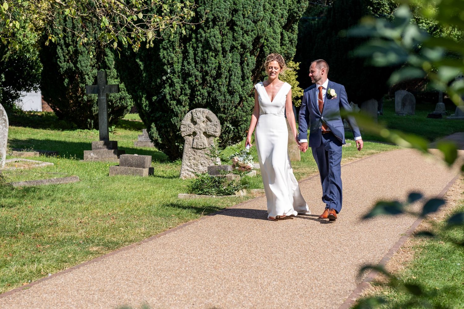 Bride and groom walking hand in hand along a sunlit church path surrounded by lush greenery and gravestones.