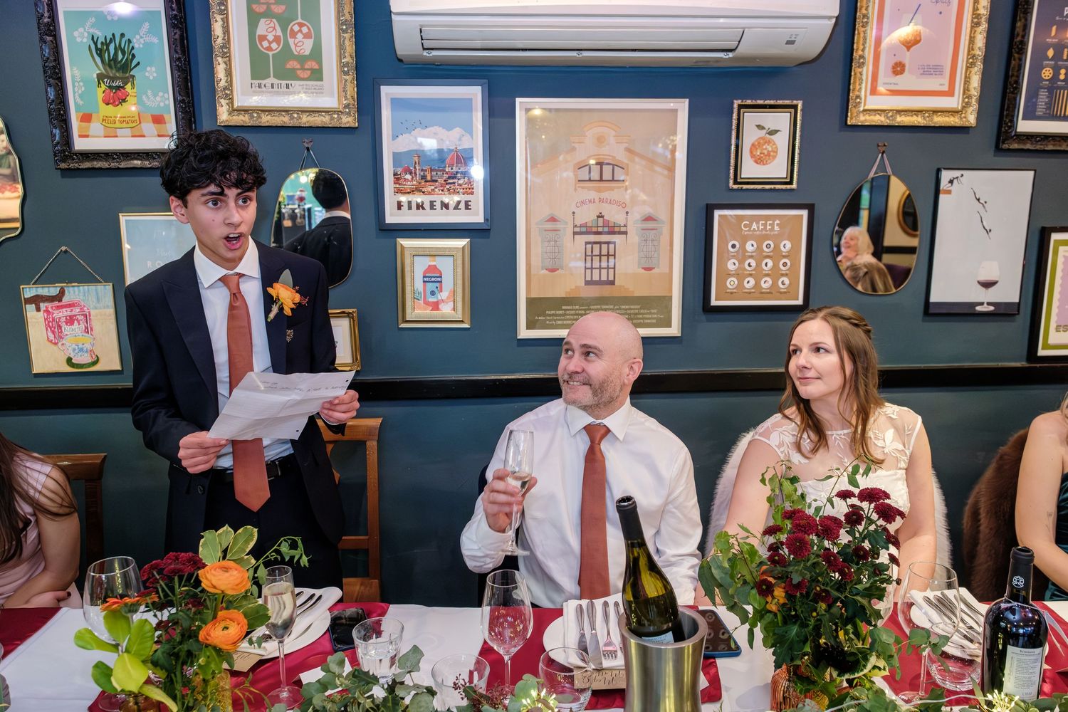 Young man reads speech at wedding reception dinner table with bride and groom seated beside him.