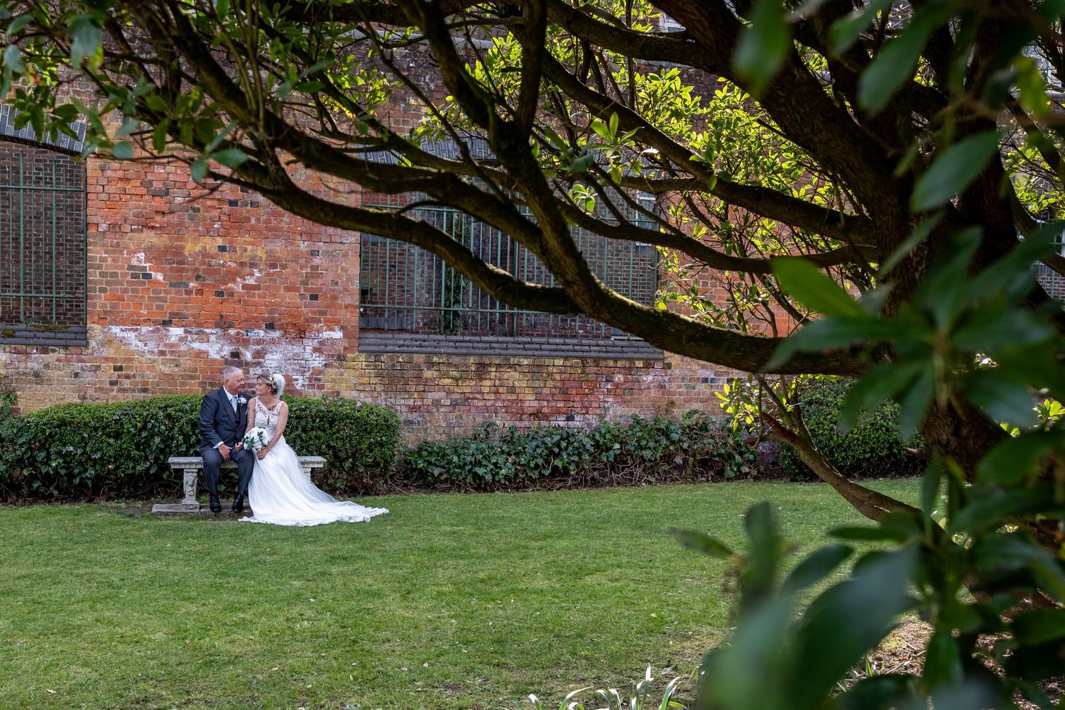 Bride and groom sitting together in a lush garden beside a historic brick building, framed by overhanging tree branches.