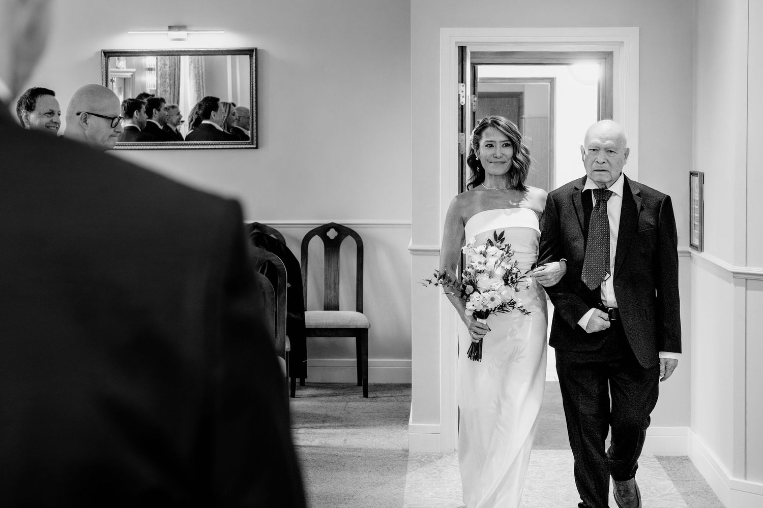Black and white photo of a bride holding a bouquet walking down the aisle with an older gentleman at a wedding ceremony.