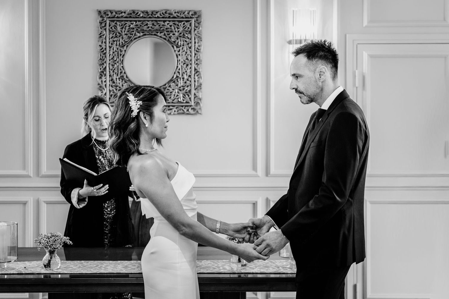 Black and white wedding ceremony photo of bride and groom holding hands before an officiant in an elegant room.