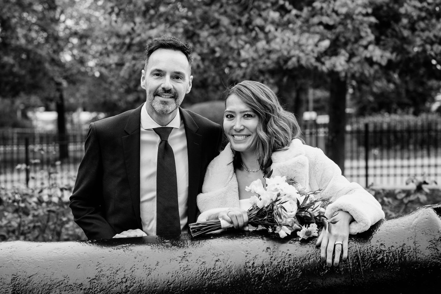 Black and white wedding photo of smiling bride and groom leaning on stone wall outdoors with trees in background.