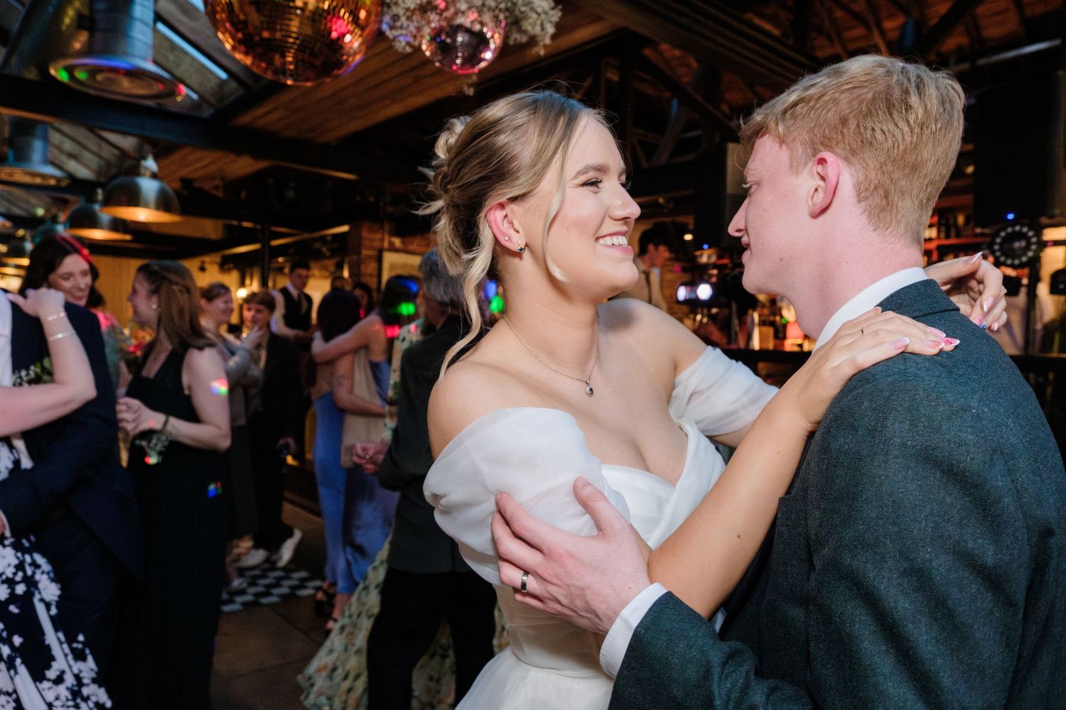 Bride and groom share first dance at wedding reception, surrounded by guests on dance floor with disco ball lighting.